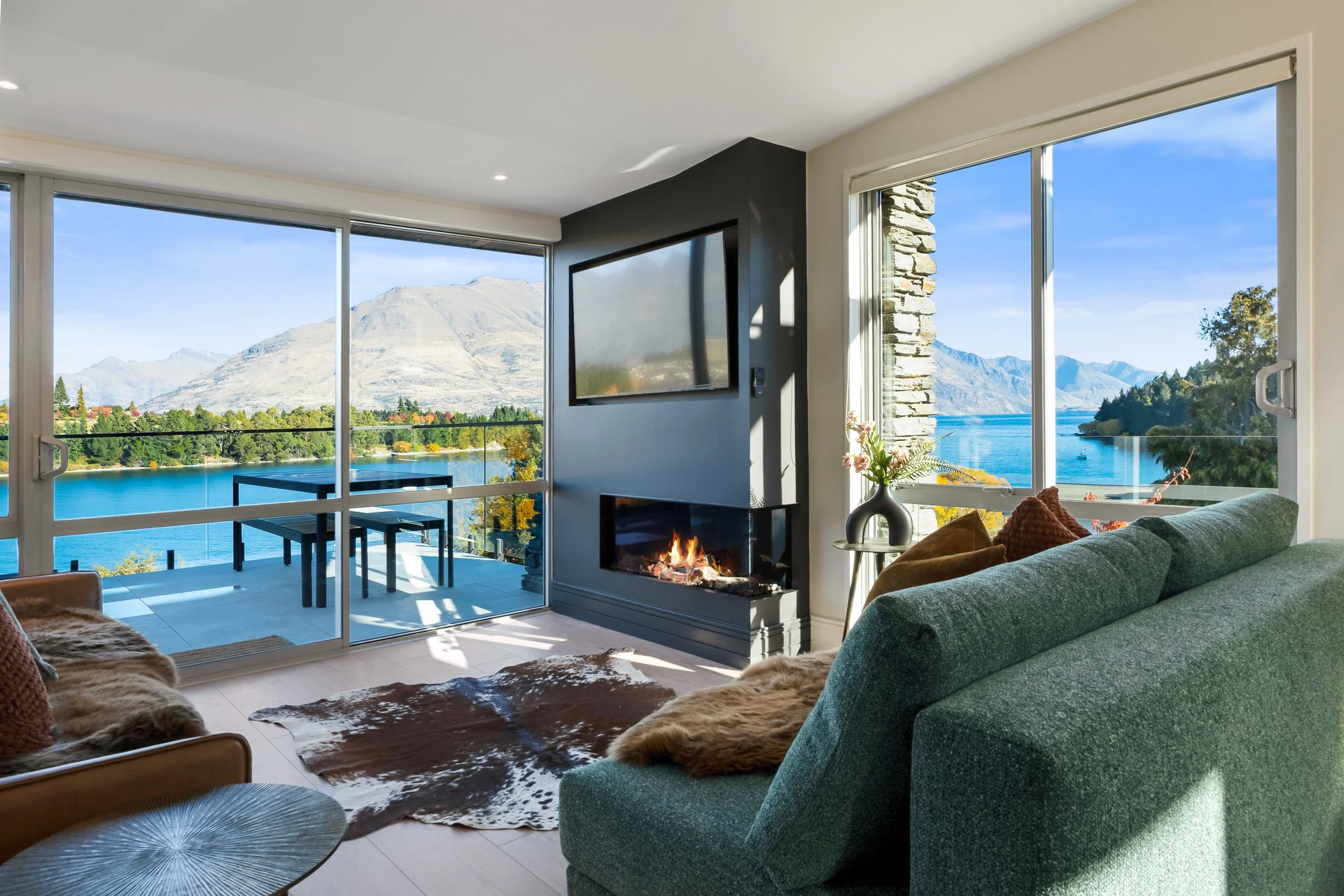 Living room with large windows showing mountains and a lake, fireplace, sofa with pillows, decorative plants, and a balcony table.