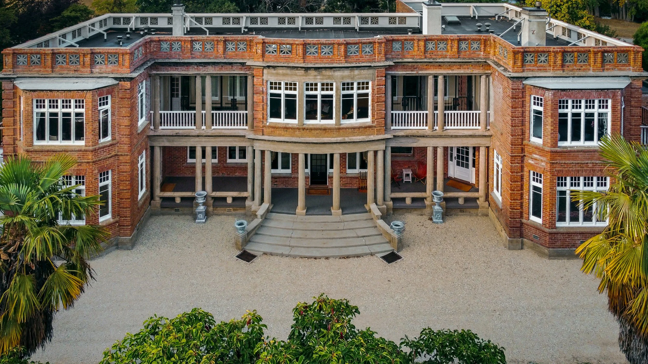 A large brick house with a curved porch, multiple large windows, and a decorative brick railing on the roof. The house has a staircase leading up to the front entrance and is surrounded by greenery and palm trees.