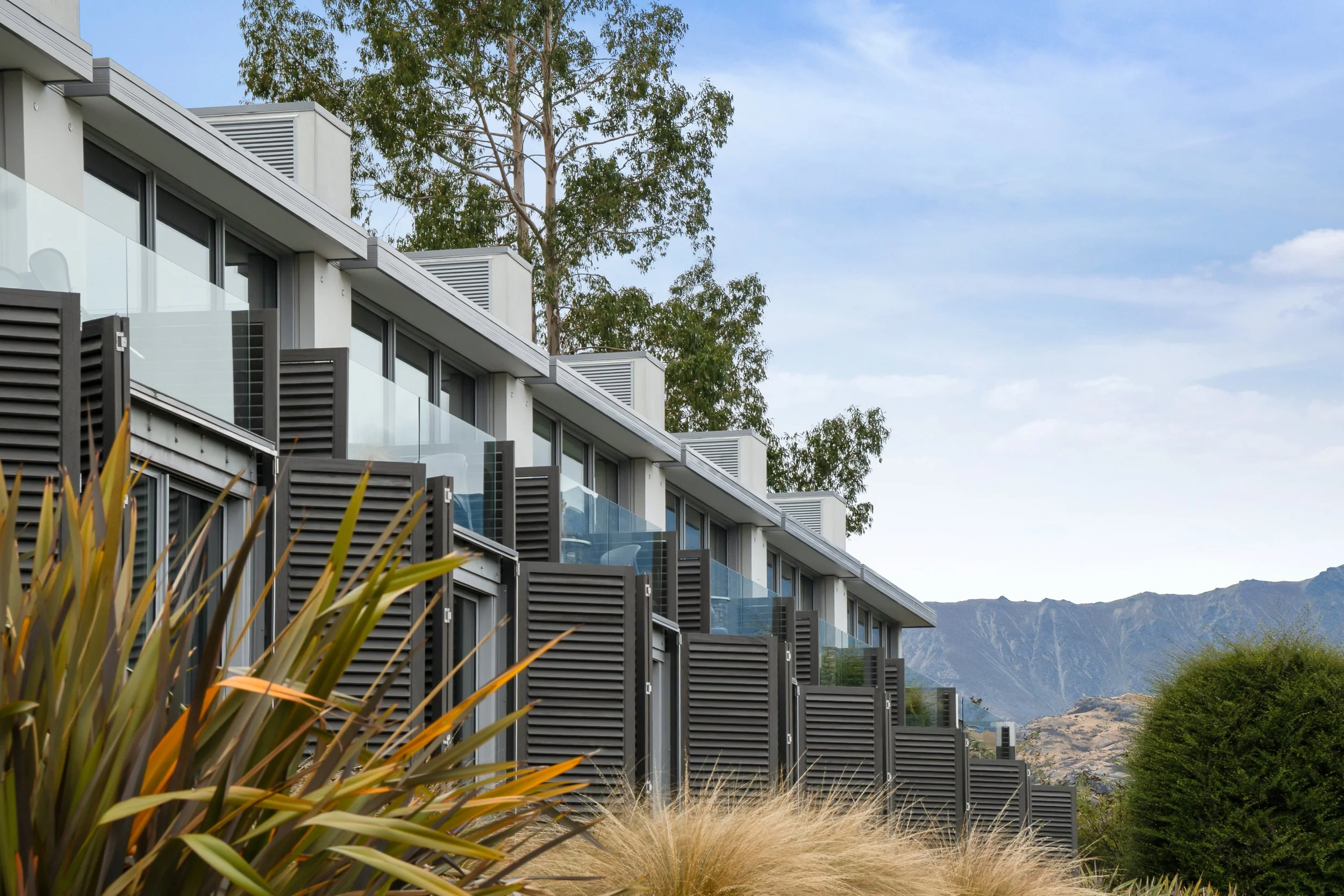 Modern multi-story apartment building with terraces, black shutters, glass railings, overlooking mountains and blue sky.