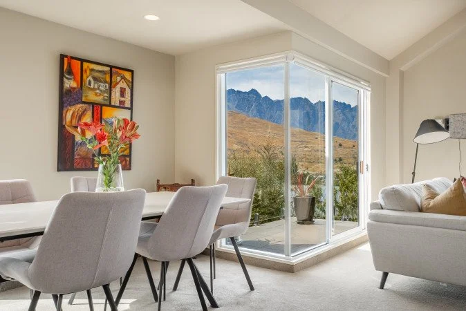 Living and dining area with large glass sliding door leading to a balcony with mountain scenery in the background; modern decor with a white table, gray chairs, a sofa, a floor lamp, and colorful artwork on the wall.