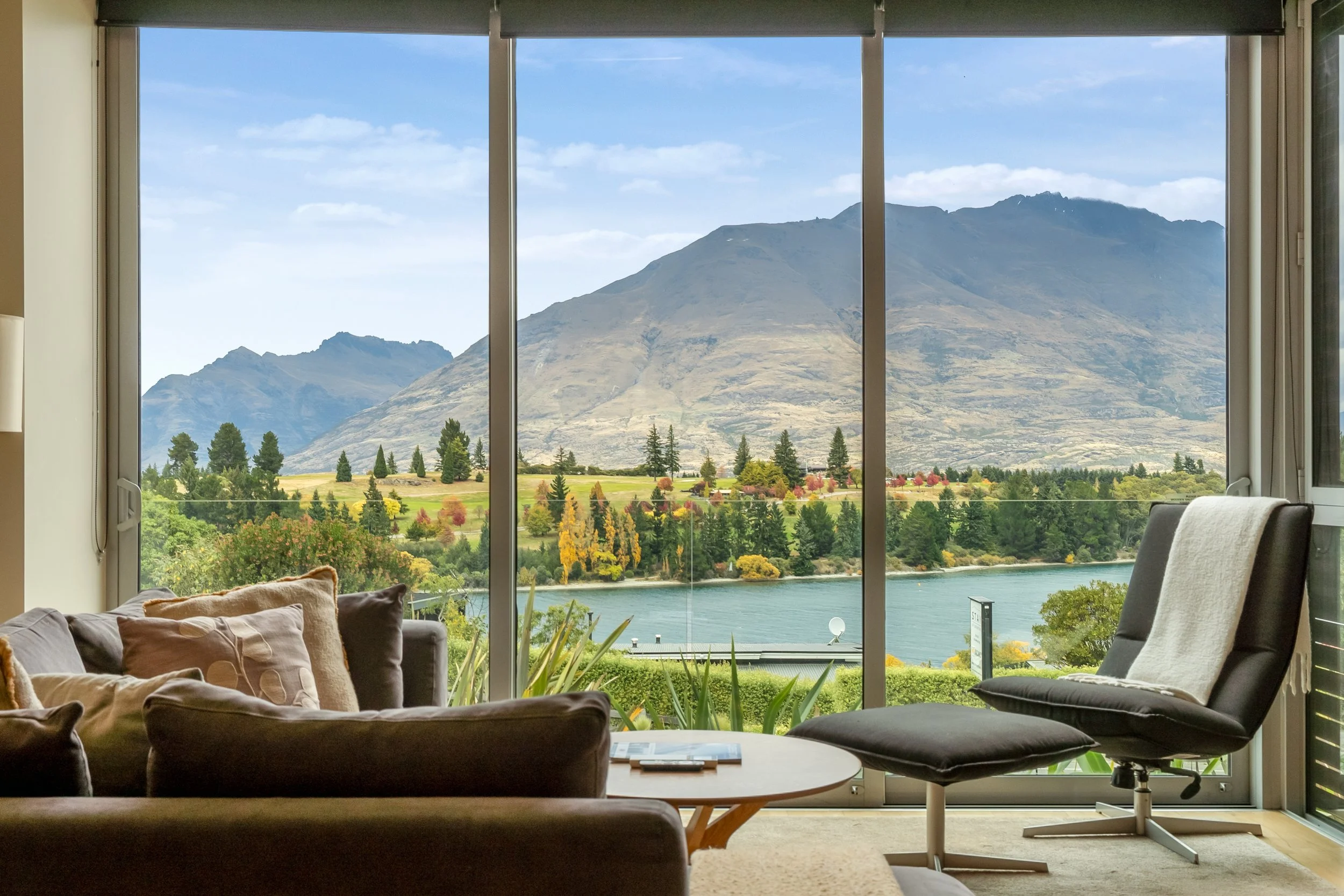 Living room with a large window view of mountains, trees, and a body of water. Furniture includes a brown sofa with pillows, a white chair, and a small table.