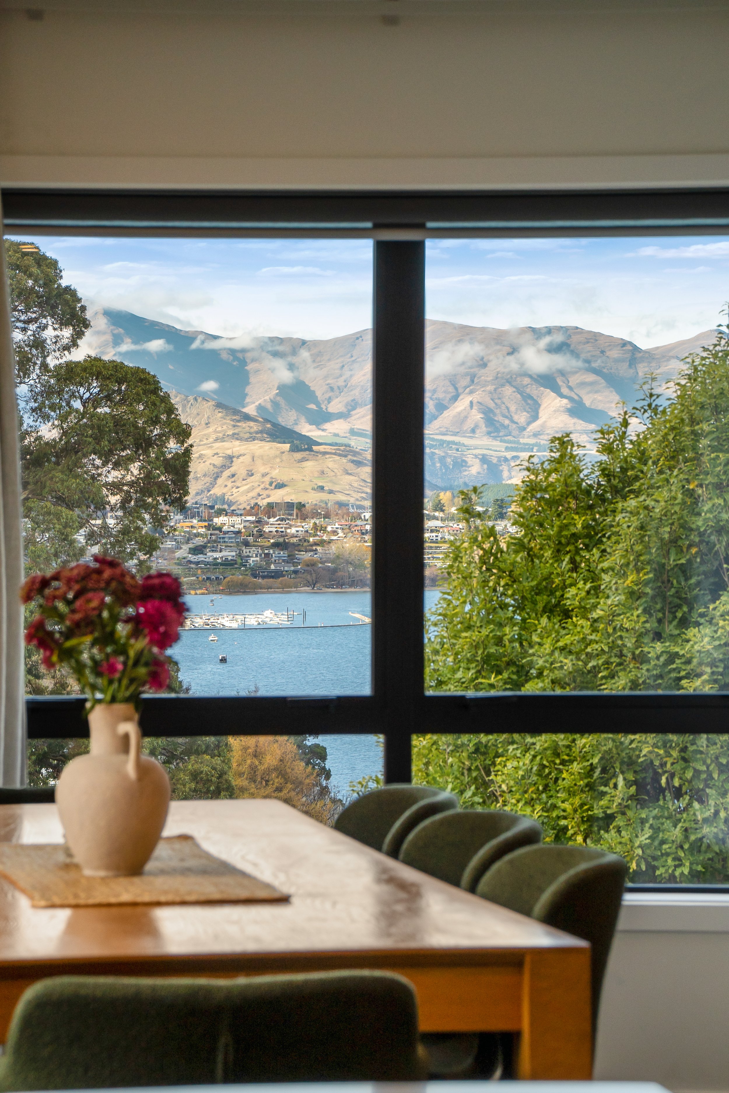 A dining table with a beige vase of pink flowers, overlooking a window with a scenic view of mountains, trees, a lake, and a town.