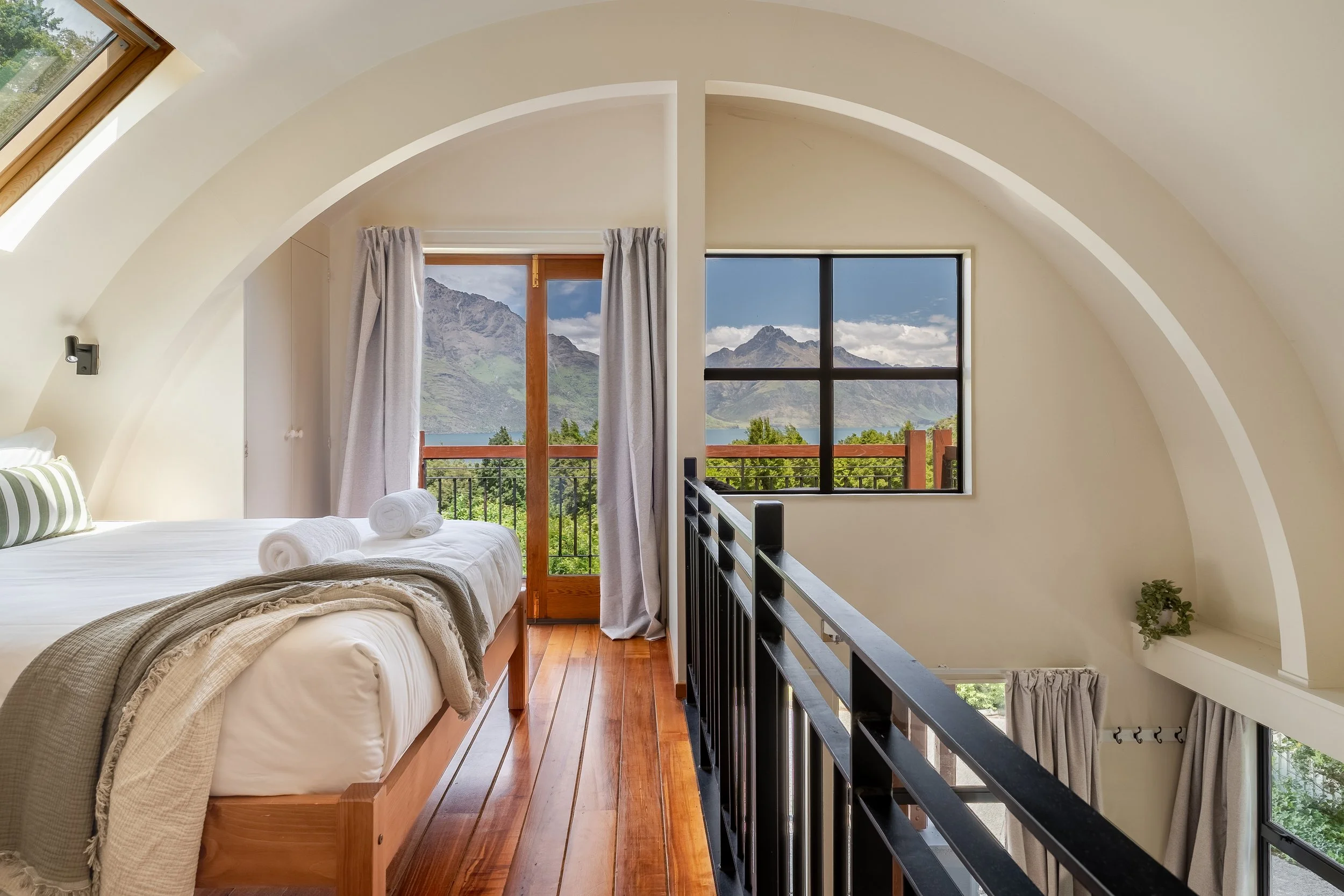 A cozy bedroom with a wooden bed, white bedding, and rolled towels, overlooking scenic mountains and greenery through large windows and a sliding glass door, with natural light and neutral decor.