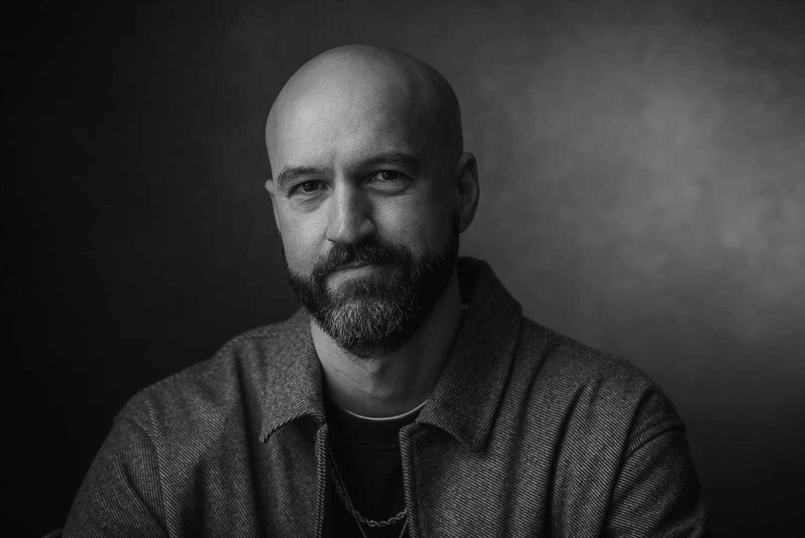 Black and white portrait of a bald man with a beard, wearing a jacket and a chain necklace, looking at the camera against a dark background.
