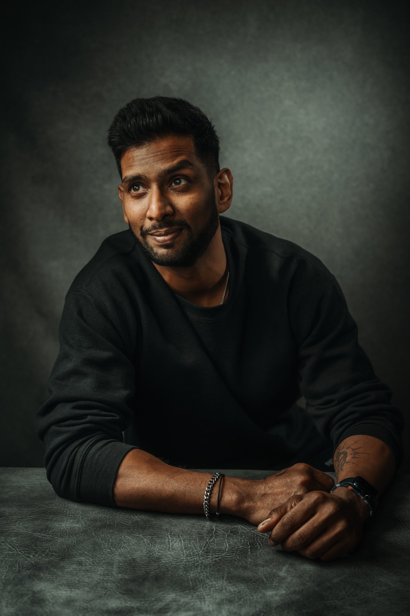A man sitting at a table against a dark background, wearing a black sweatshirt, with short black hair, a beard, and tattoos on his left wrist. He is looking slightly to the side with a slight smile.