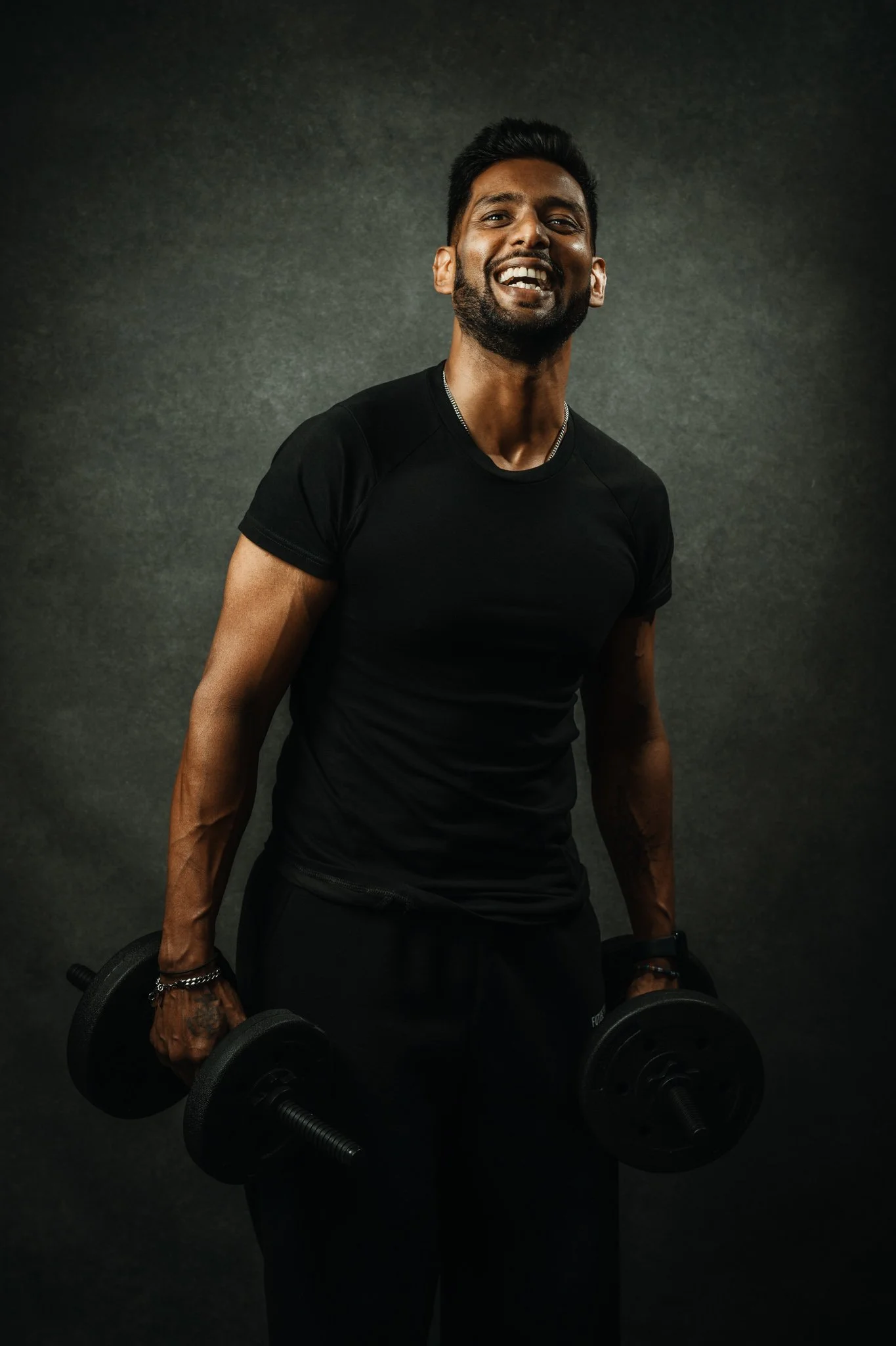 A young man with a beard and dark hair smiling while holding dumbbells in both hands, wearing a black t-shirt and black pants, against a dark background.