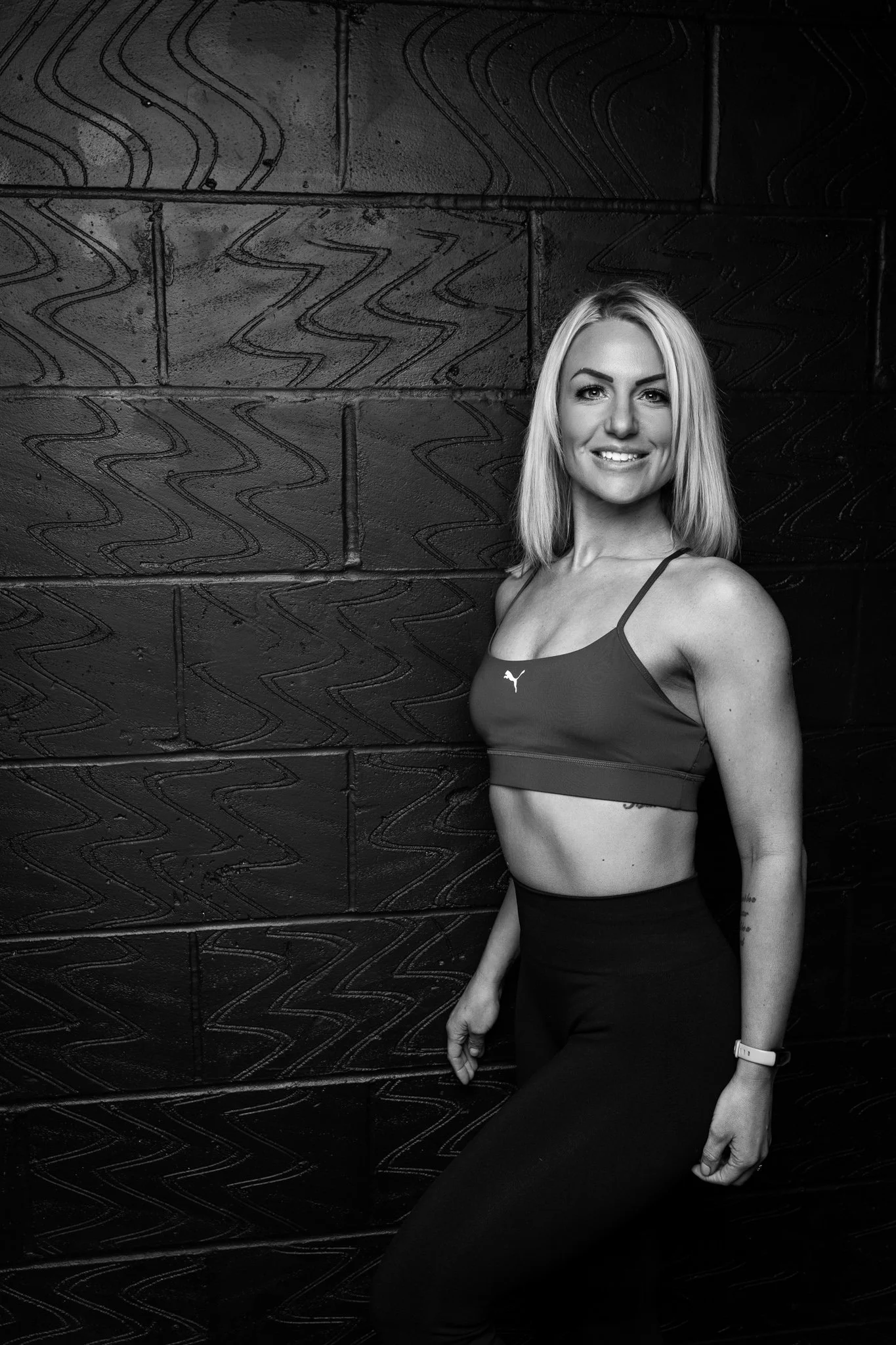 Black and white photo of a smiling woman with blonde hair in workout attire, standing against a textured brick wall.