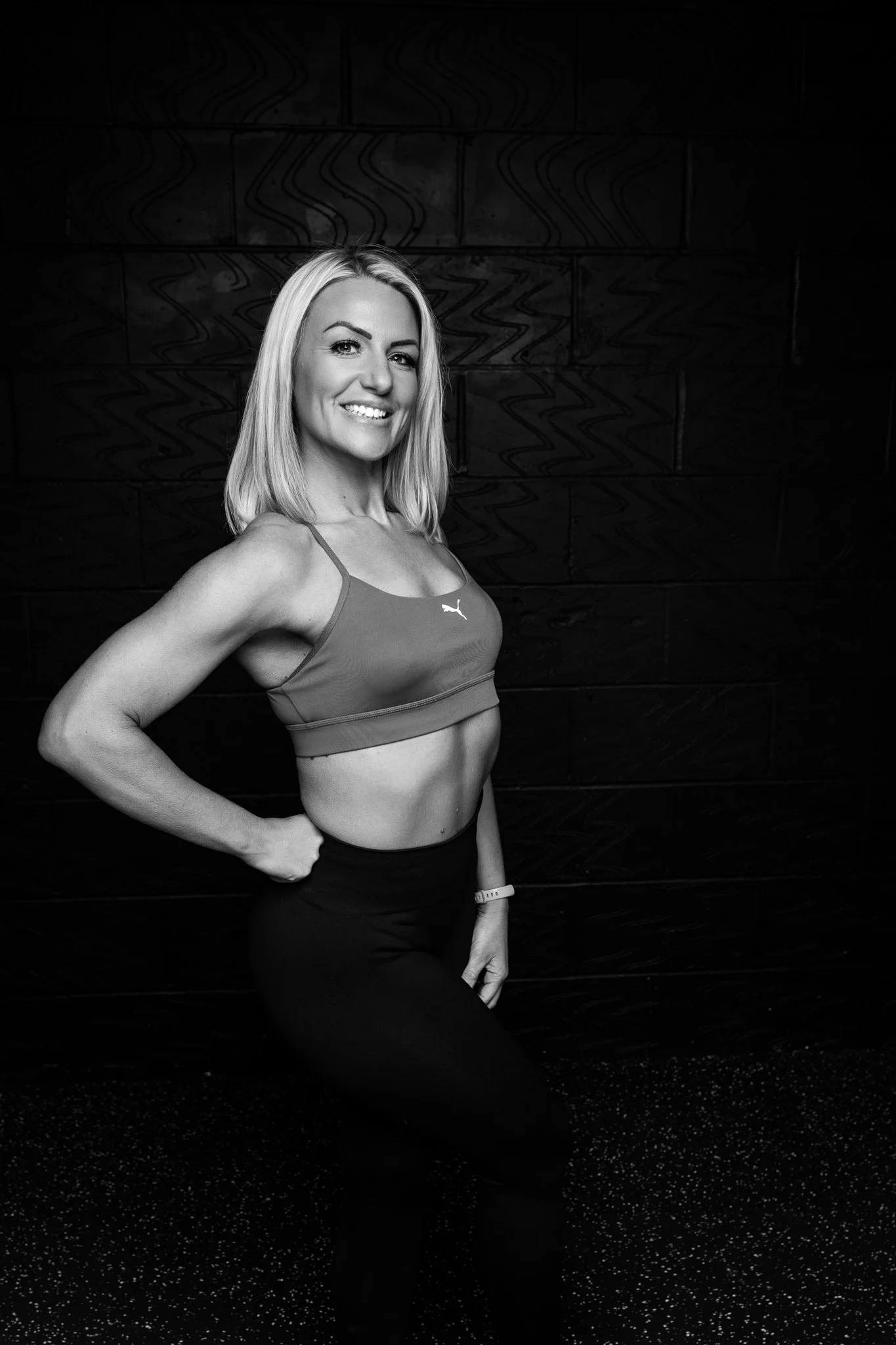 A woman with blonde hair in athletic wear, smiling and posing with her hand on her hip against a dark textured background.