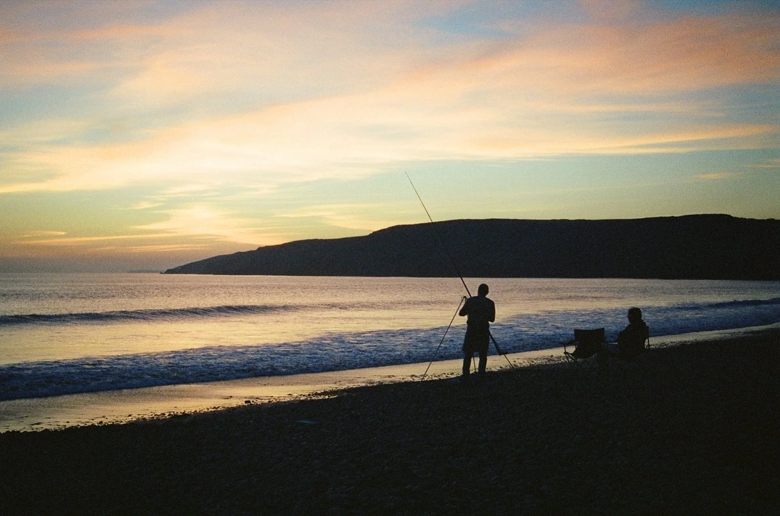Silhouettes of two people on a beach during sunset, one standing with a fishing rod and the other sitting in a chair, with calm ocean waves and a distant landform in the background.