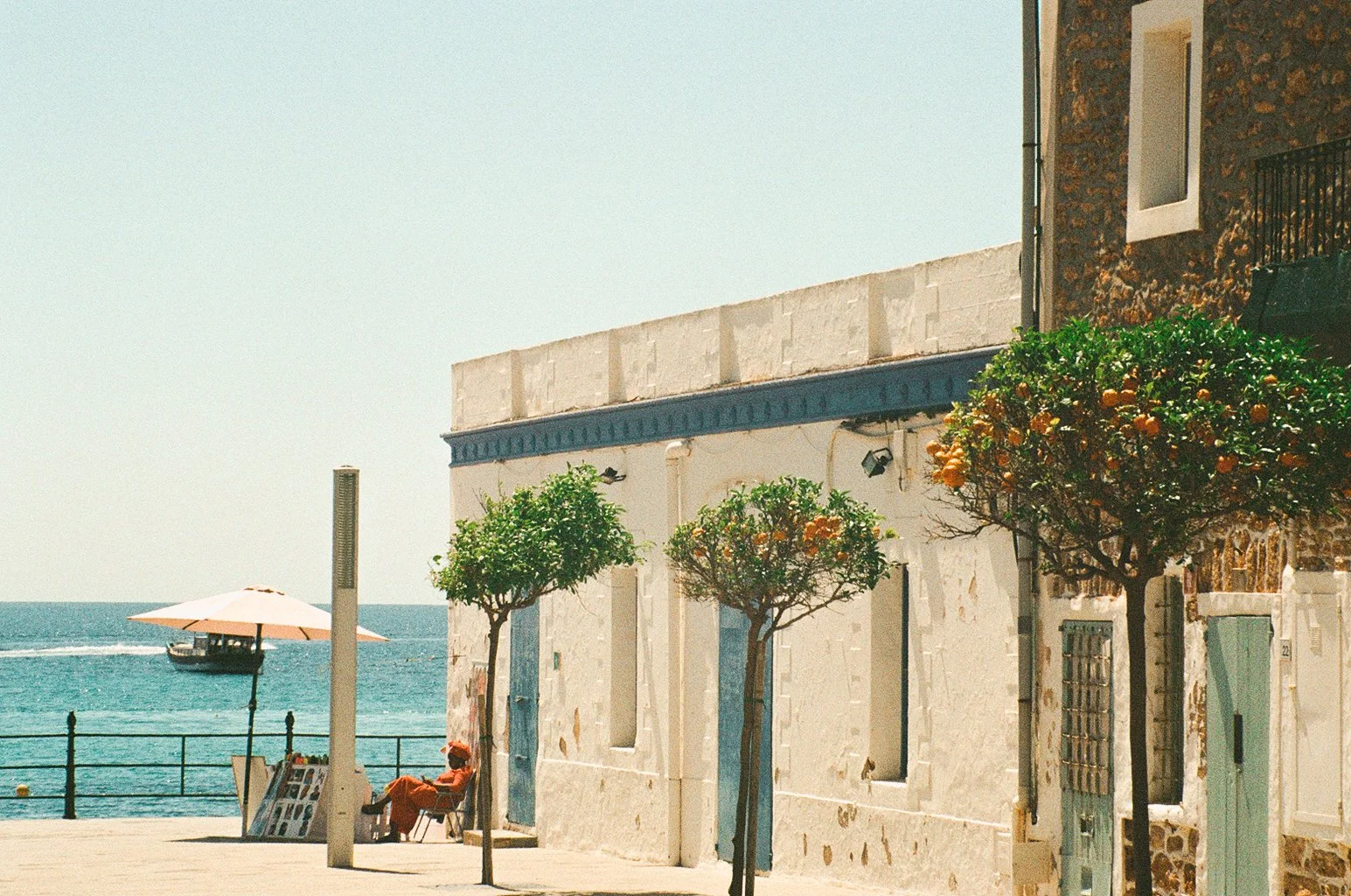 A seaside scene with orange trees, a person sitting on a chair under an umbrella, a boat on the water, and buildings facing the street.