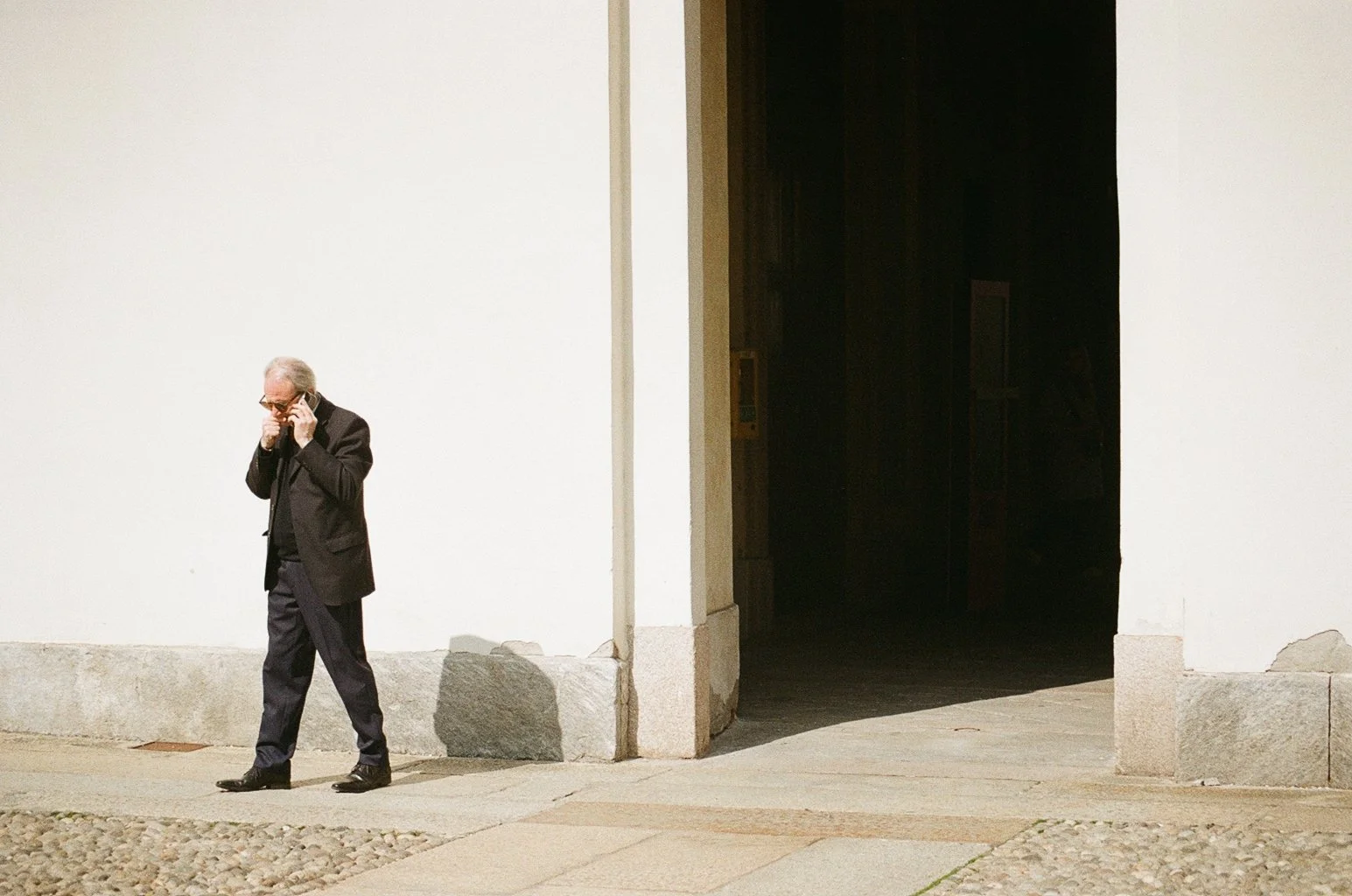 An older man in a black suit and glasses is talking on his cellphone while walking along a sidewalk outside a white building with a dark entrance.