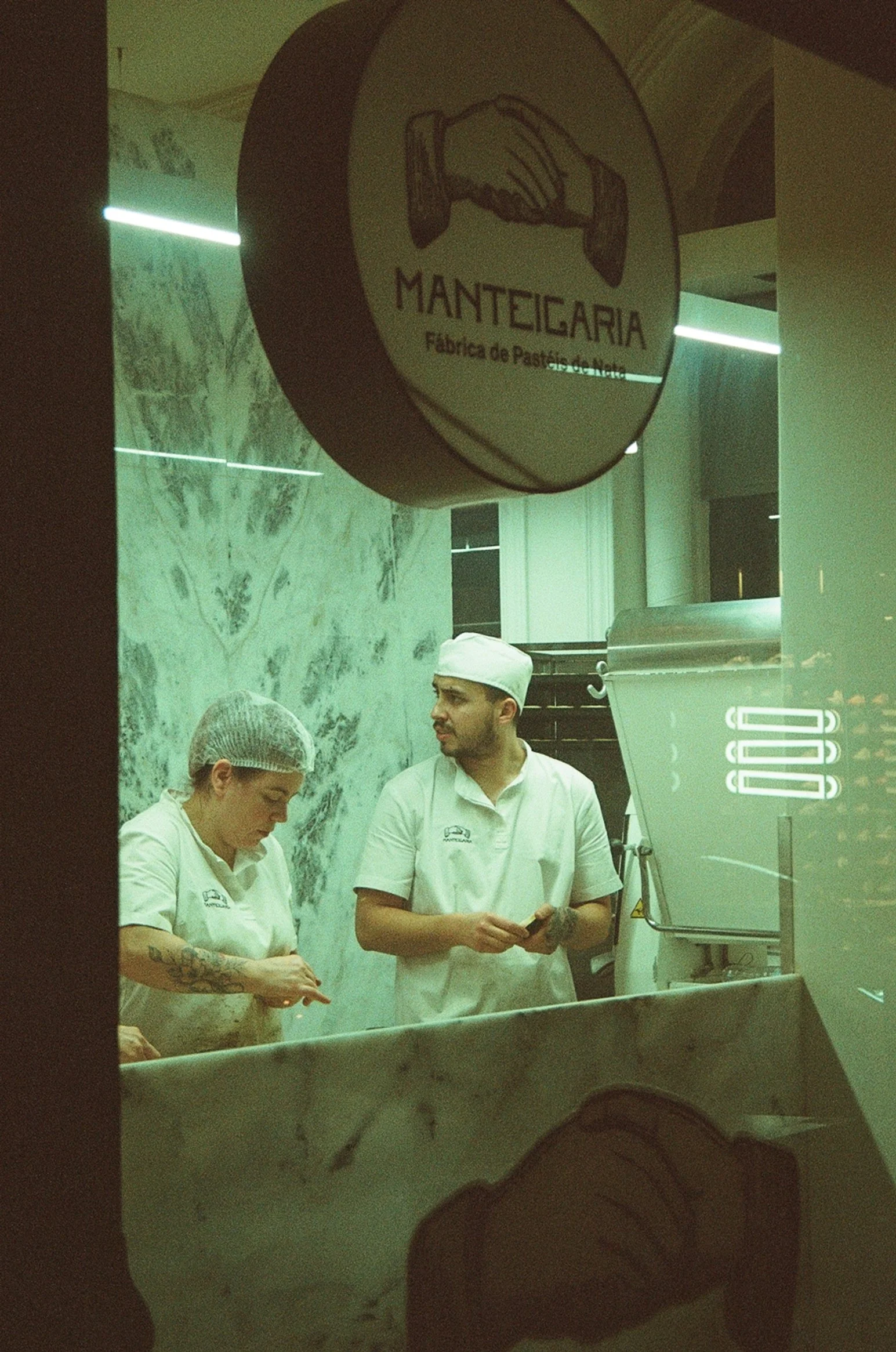 Inside a pastry shop named Manteigaria, with two staff members wearing white uniforms and hairnets. One staff member has tattoos on their arm, and a male staff member in the middle is making a pastel de nata. There is a marble wall in the background 