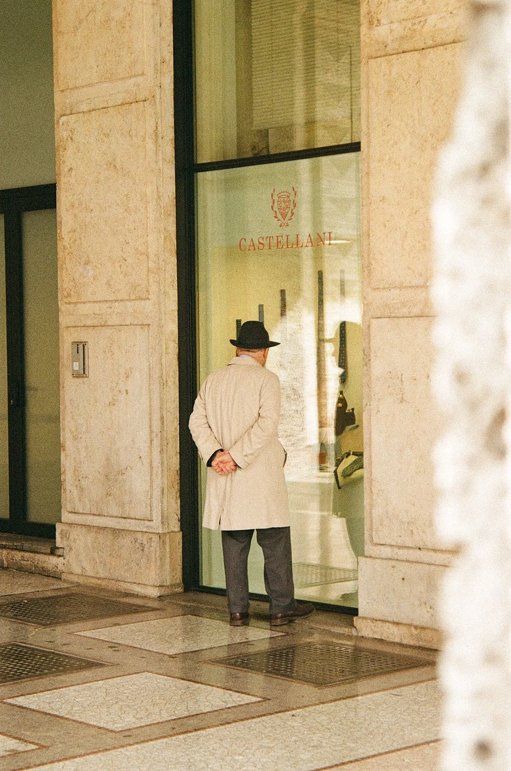 A man in a beige coat and black hat stands outside a store, window shopping, in a city setting with stone walls and tiled pavement.