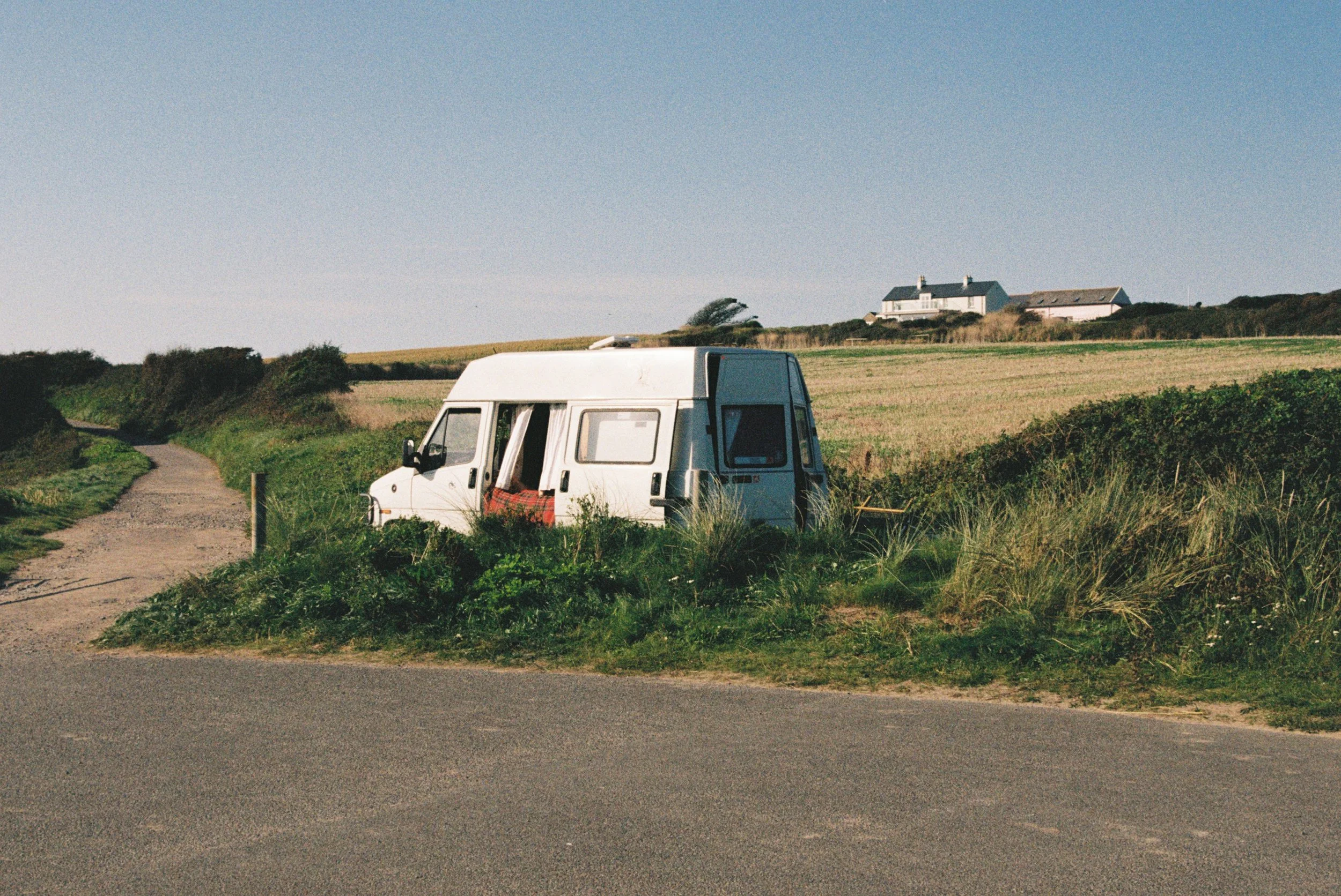 A small white camper van parked on grassy area beside a gravel path with a house on a hill in the background and clear blue sky.