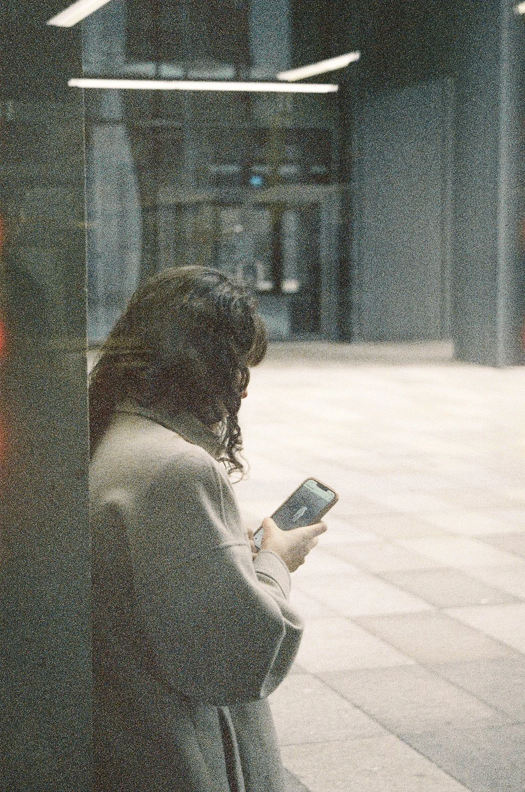 A woman with curly hair leaning against a wall in an urban setting, looking at her smartphone.