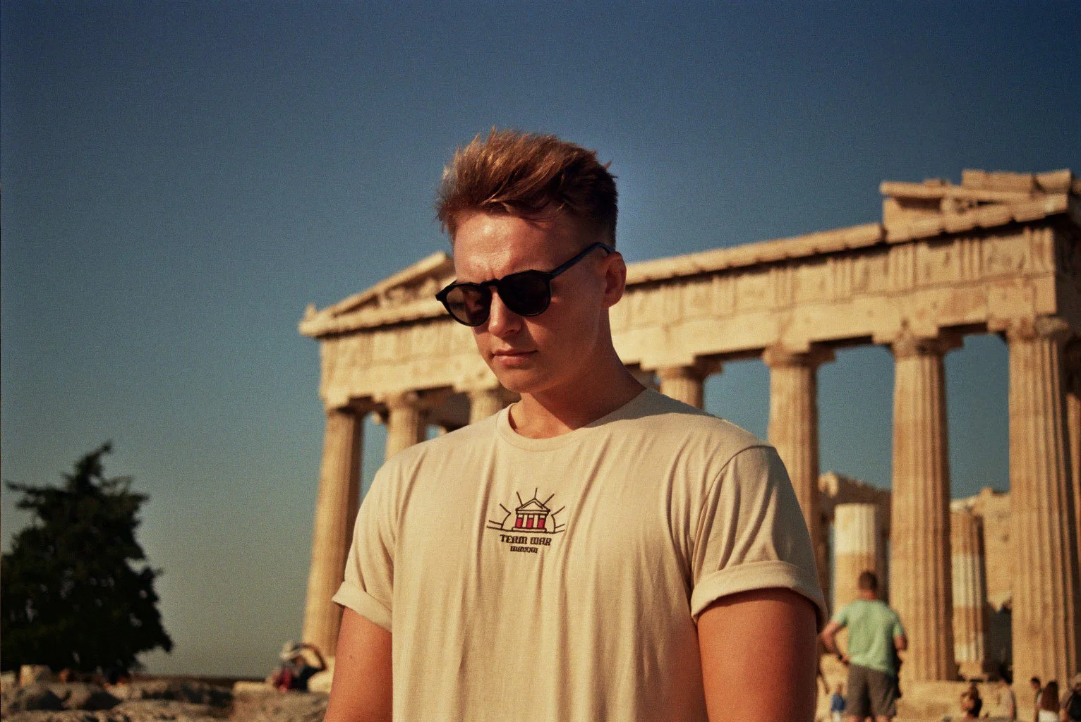 Dewi Thomas, the branding specialist and designer wearing sunglasses and a beige t-shirt stands in front of the ancient Parthenon temple in Greece during sunset.