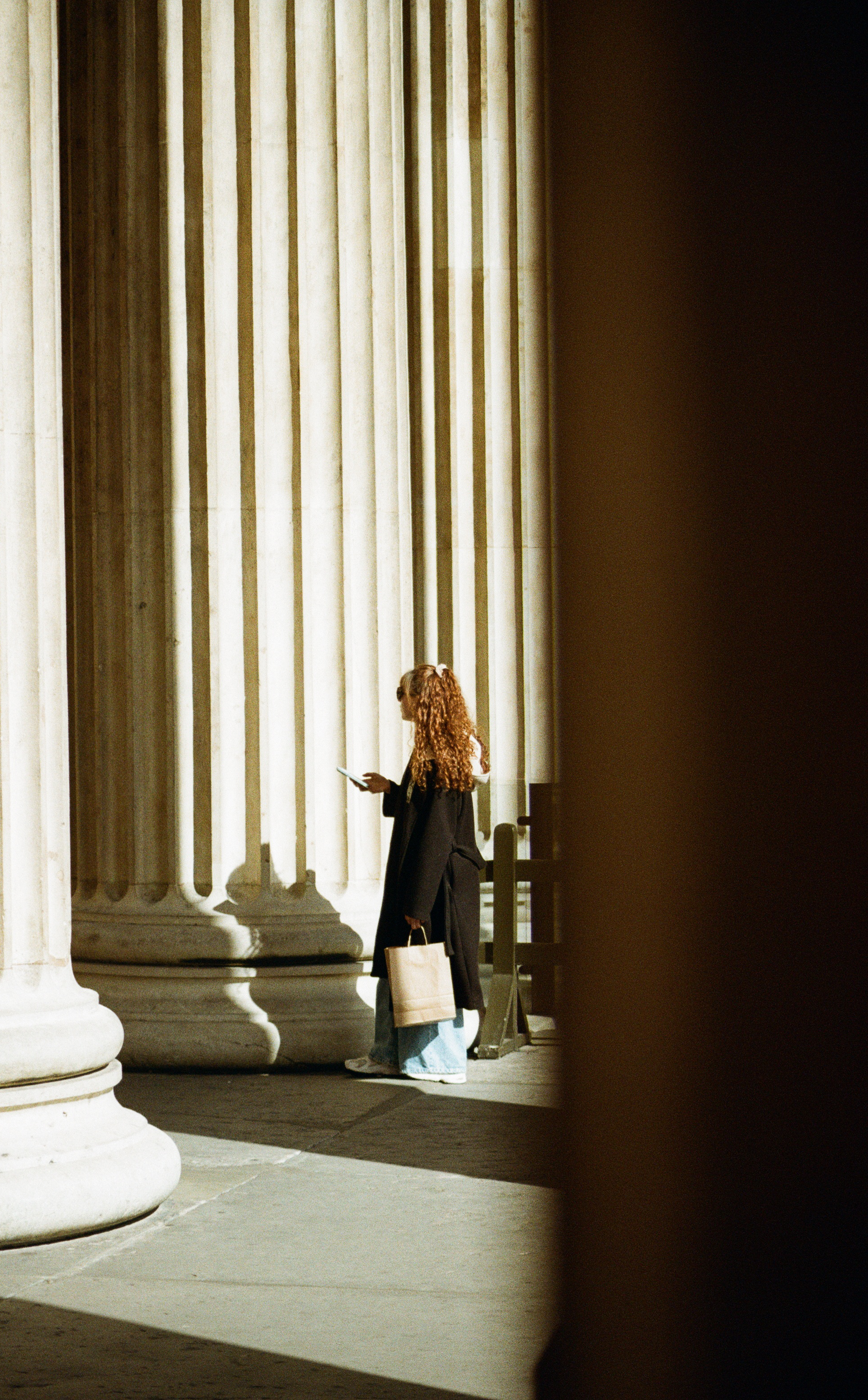 A woman with curly hair wearing a black coat and light blue jeans, standing near large white columns of a classical building (British Museum London), holding a tote bag and looking at her phone.