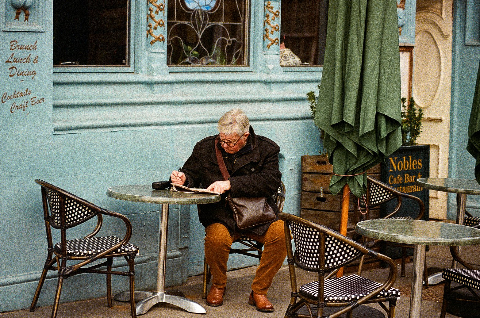 An older man with short gray hair and glasses sitting alone at an outdoor cafe table, writing in a notebook, with a black sachel bag on his lap. The cafe has blue painted walls, a sign indicating "Nobles Cafe Bar & Restaurant," and closed umbrellas o