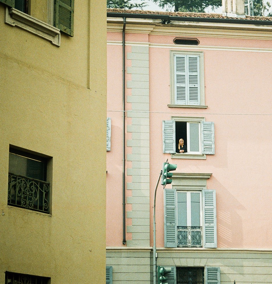 A woman looking out of a second-story window of a pink building with blue shutters in an urban area, near a traffic light.