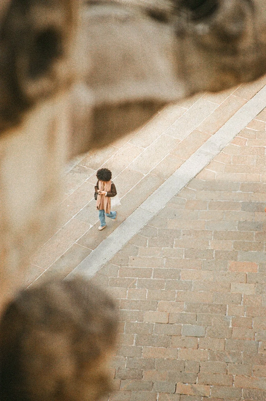 A woman with curly hair walking in Milan's Duomo square, holding a phone and wearing a scarf, in an overhead view.