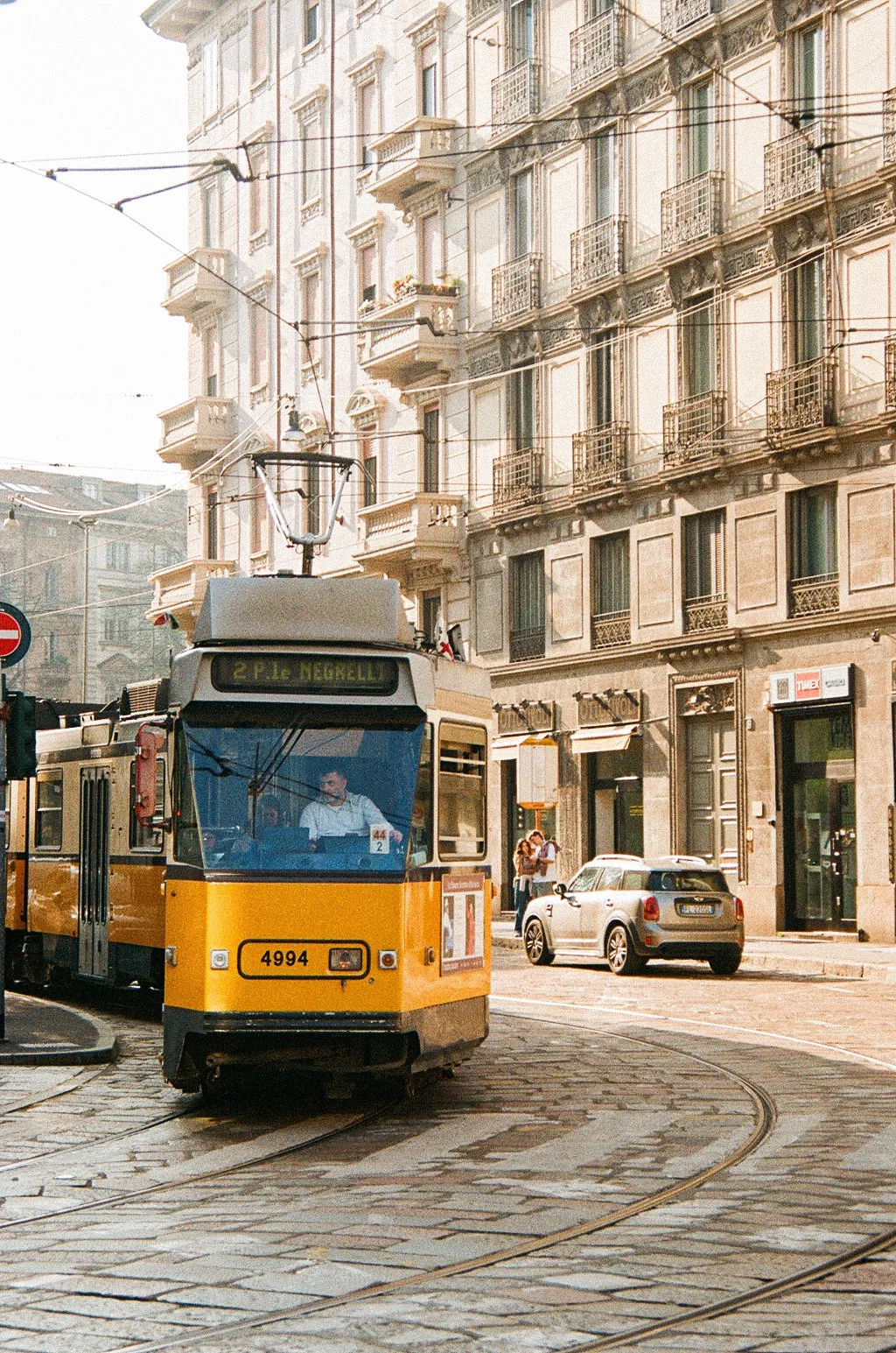 A yellow tram on cobblestone street in front of a classic European apartment building with balconies in an urban city setting (Milan), with cars and pedestrians nearby.