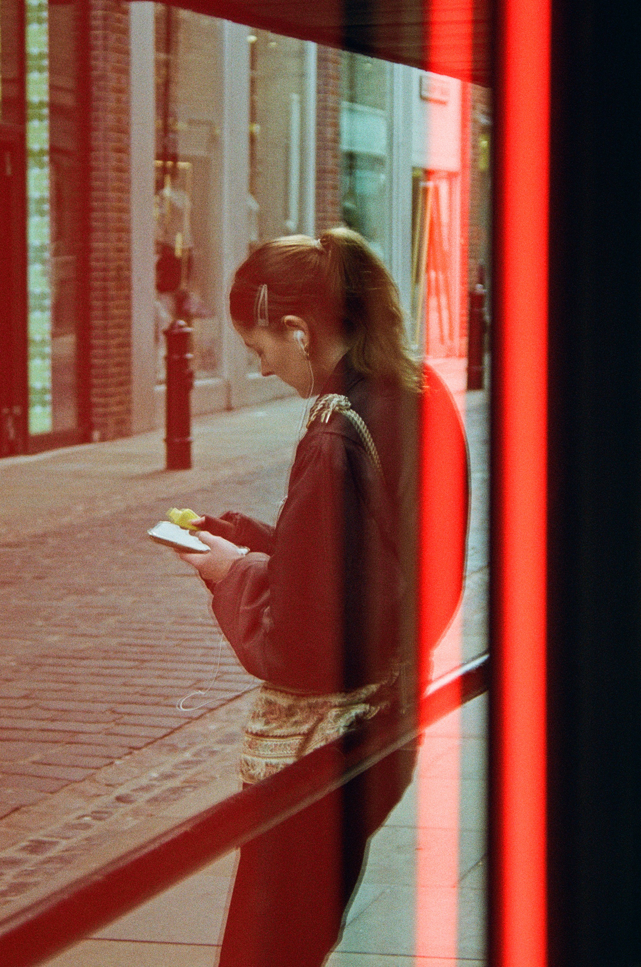 A woman with short brown hair, wearing a dark jacket is leaning against a large windowed building facade and looking at her smartphone with earphones on. The photo appears to be taken through the glass reflection with red framing.