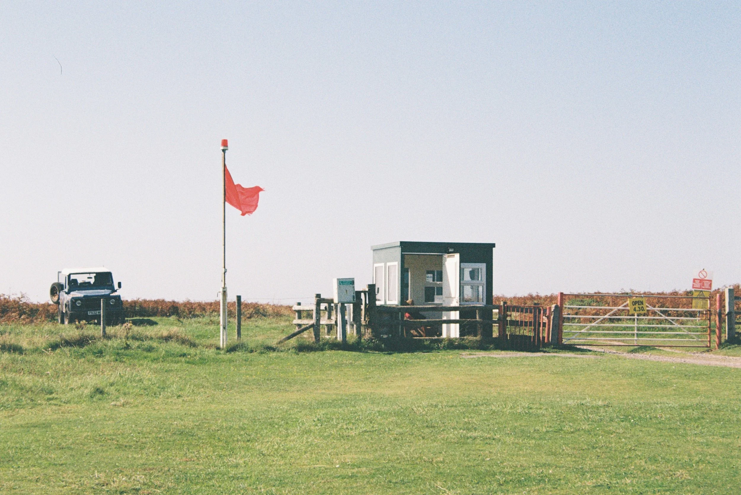 A grassy field with a small military lookout structure, a flagpole with a red flag, a vehicle, and some signage in the background under a pale blue sky.