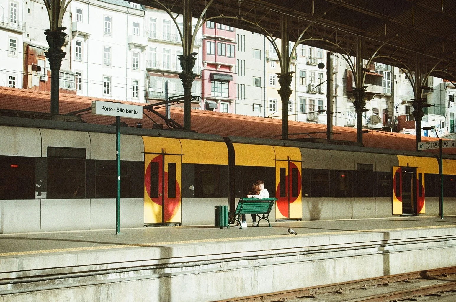 A train at the Porto - São Bento train station, with a couple sitting on a green bench and a pigeon nearby. The station features ornate iron supports and has residential buildings in the background.
