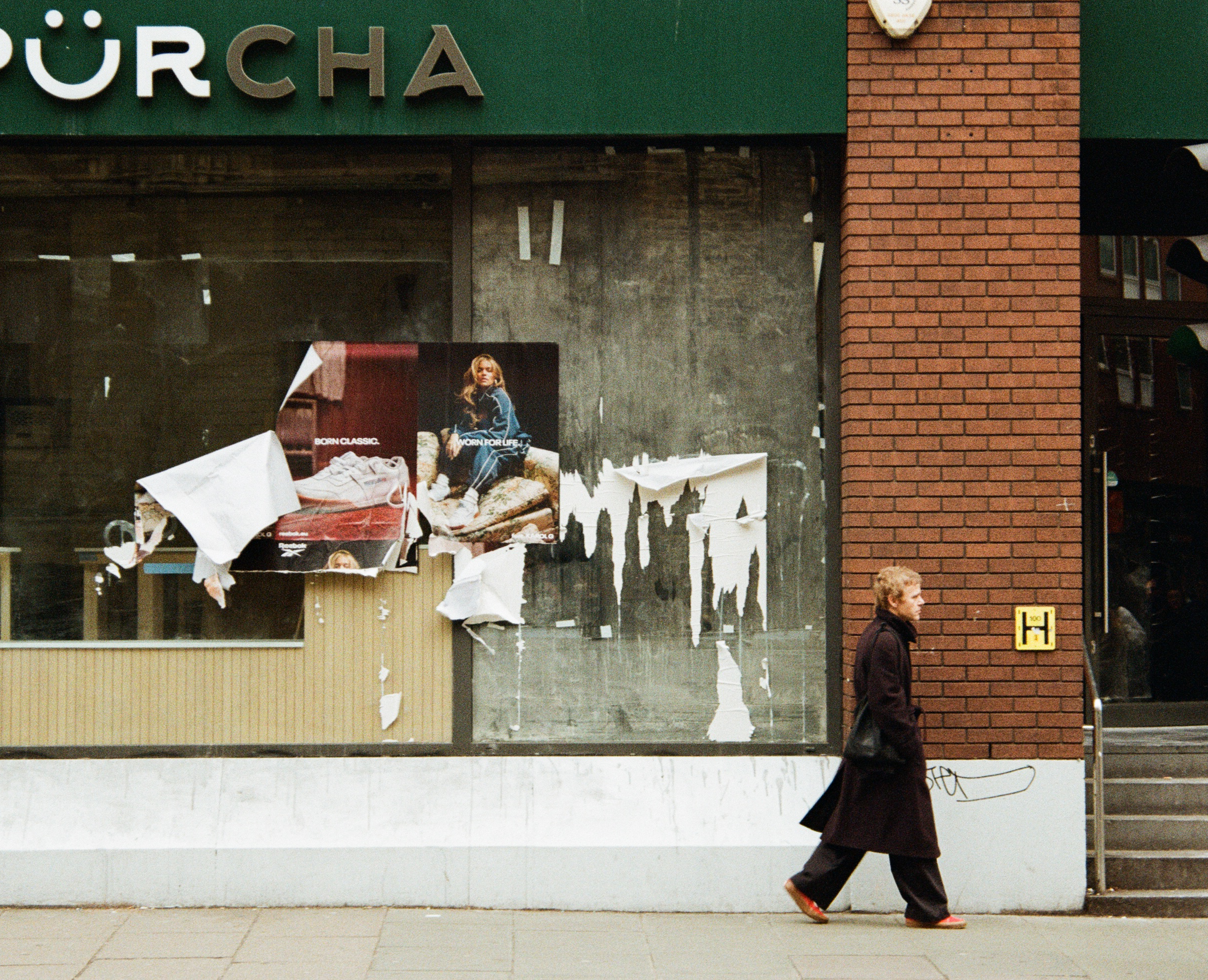 A person in a dark coat and black pants walking alonga London pavement in front of a building with a brick wall and ripped posters.