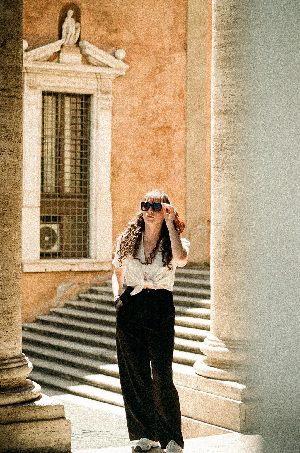 A woman with long curly hair, wearing sunglasses, a white tied blouse, and black wide-leg pants, stands between large stone pillars in Rome.