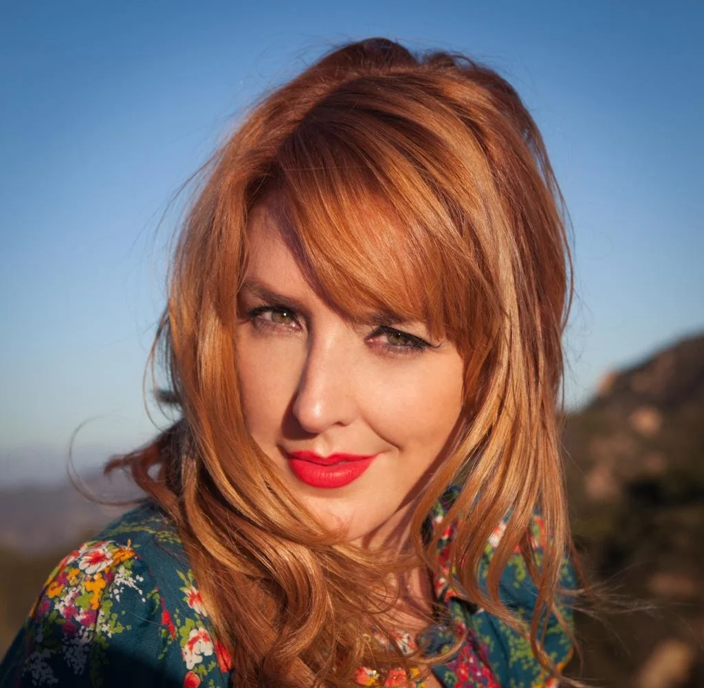 The singer from Urban Cookie Collective -  with red hair and green eyes winks at the camera outdoors, wearing a floral top with a mountain background.
