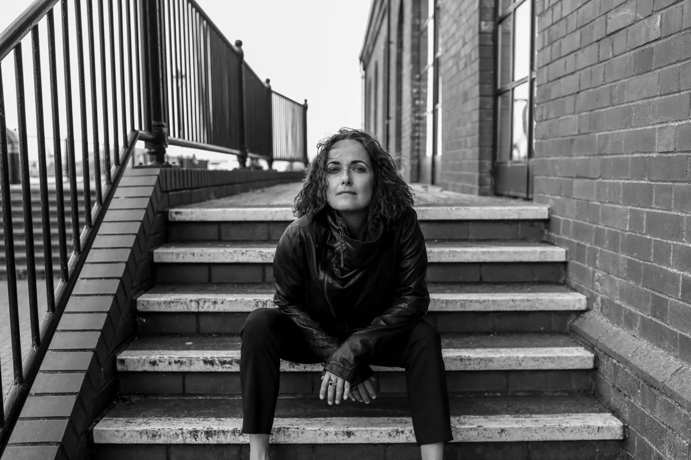 Black and white photo of a woman with curly hair sitting on outdoor stairs between brick walls, looking at the camera with a serious expression.