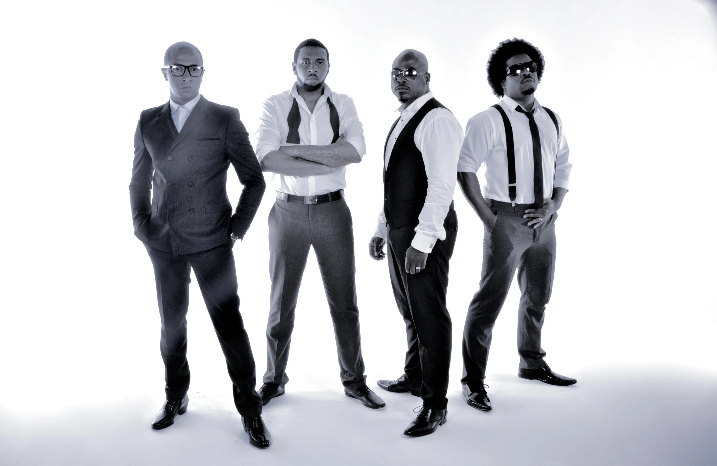 Four men dressed in black and white formal suits and shirts, standing against a plain white background.