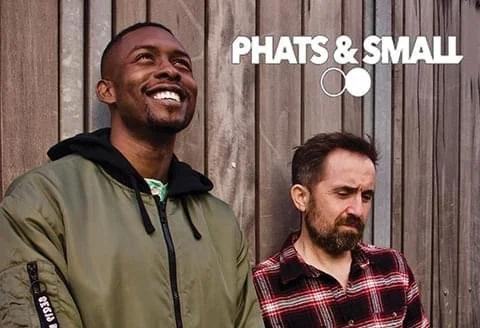Two men standing in front of a wooden fence, one Smiling and looking up, the other with a serious expression. Text reads "PHATS & SMALL".