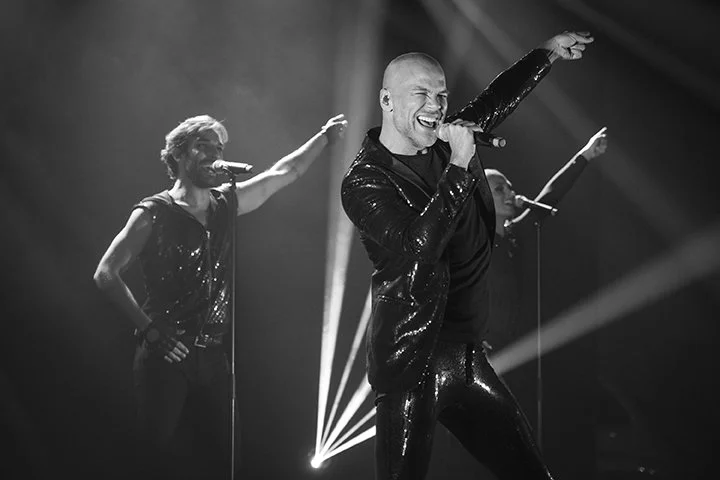 A male singer passionately performing on stage with two backup dancers, all dressed in shiny black outfits, under stage lights.