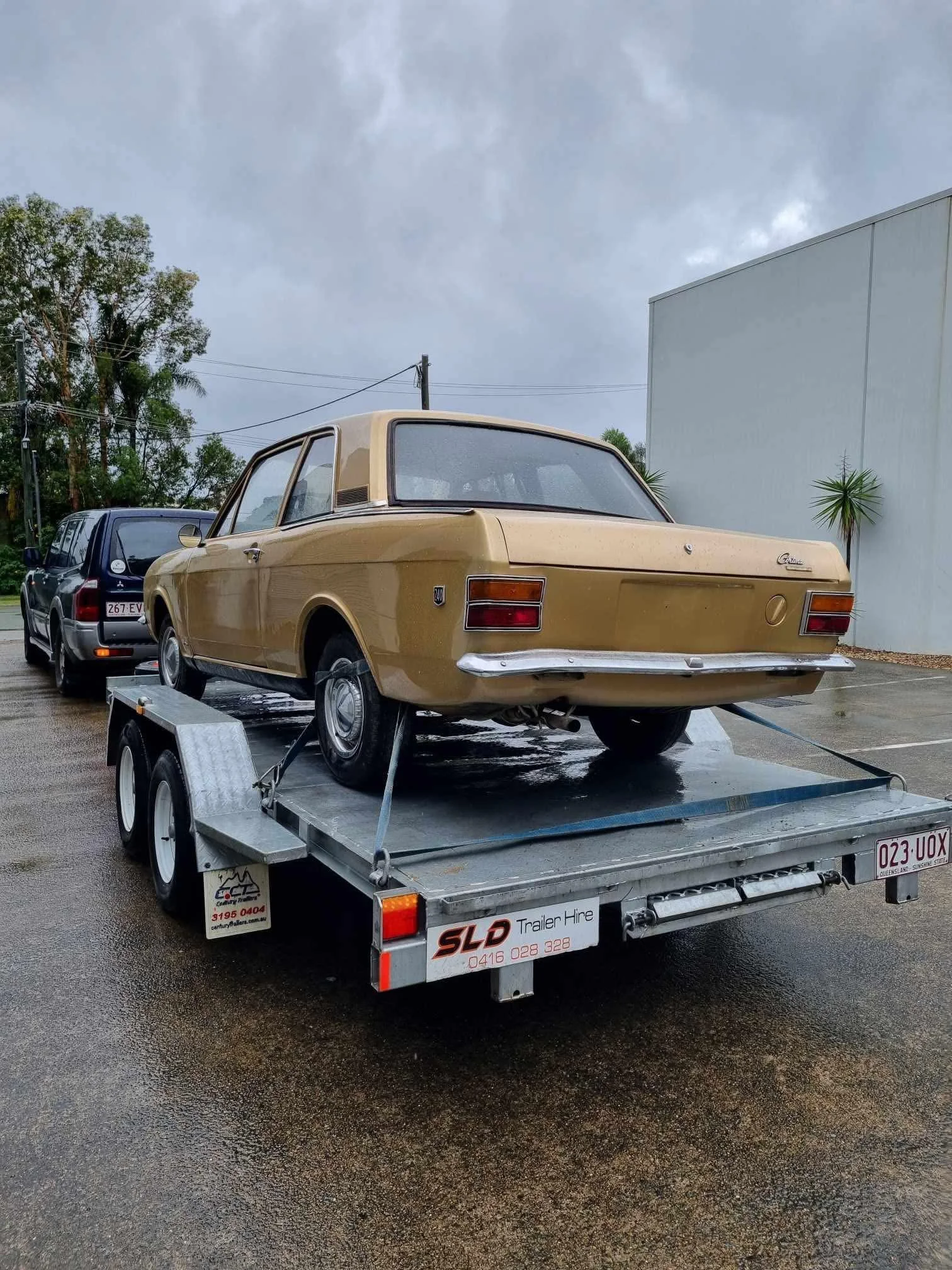 SLD Trailers and Plant Hire Gold Coast. A vintage gold car on a flatbed trailer in a parking lot on a rainy day.
