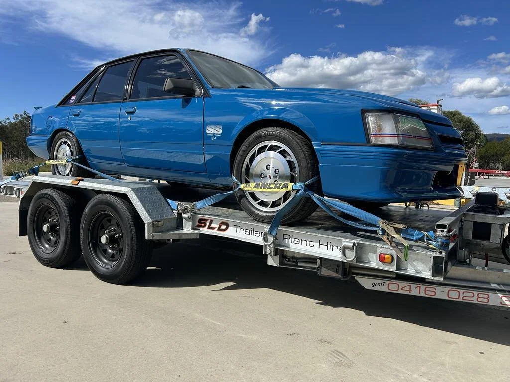 SLD Trailers and Plant Hire Gold Coast. A vintage blue sedan car secured on a flatbed trailer for transport, outdoors under a partly cloudy sky.