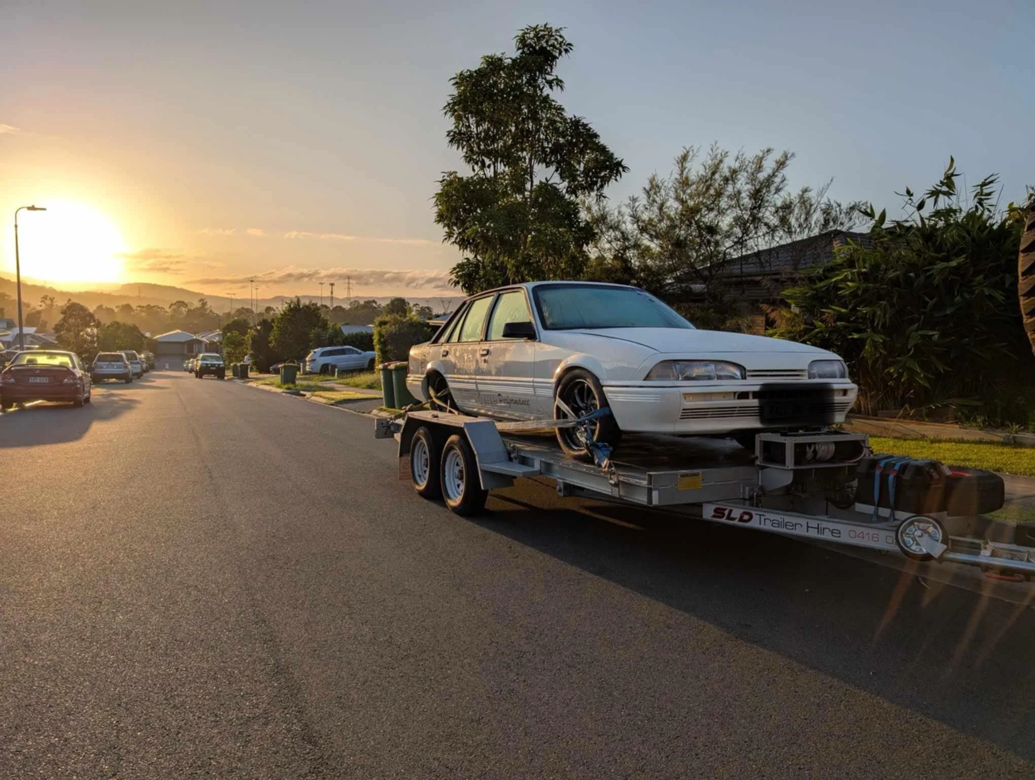 SLD Trailers and Plant Hire Gold Coast. A white car is on a trailer parked on a suburban street during sunset, with other cars and houses visible in the background.