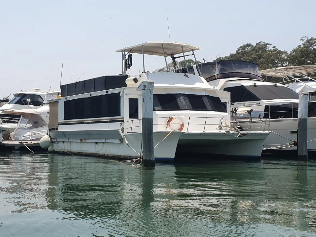 A white houseboat moored at a marina, surrounded by other boats and yachts, with trees in the background.