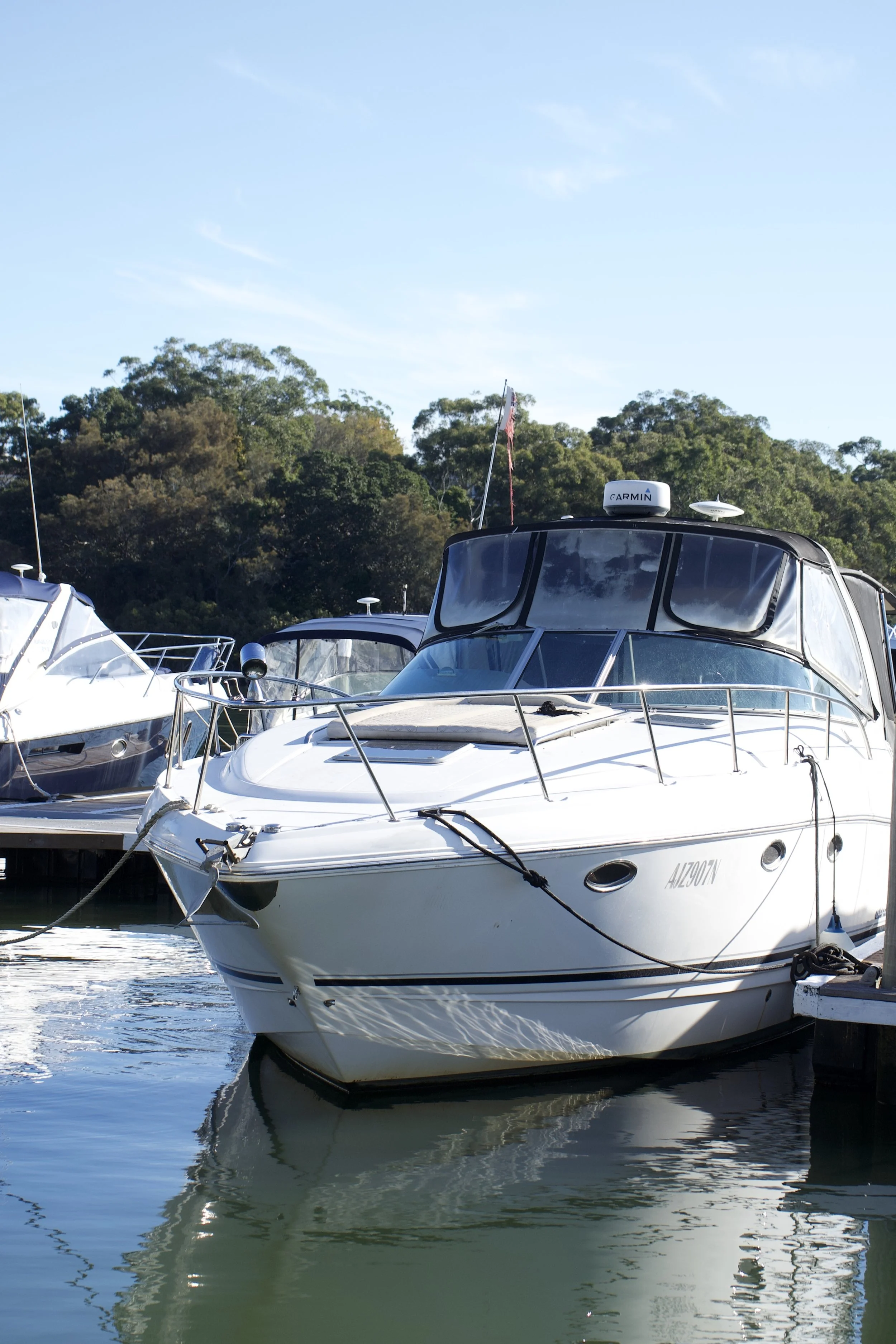 A white motor yacht docked at a marina with a reflection in the water, surrounded by other boats and green trees in the background under a clear blue sky.