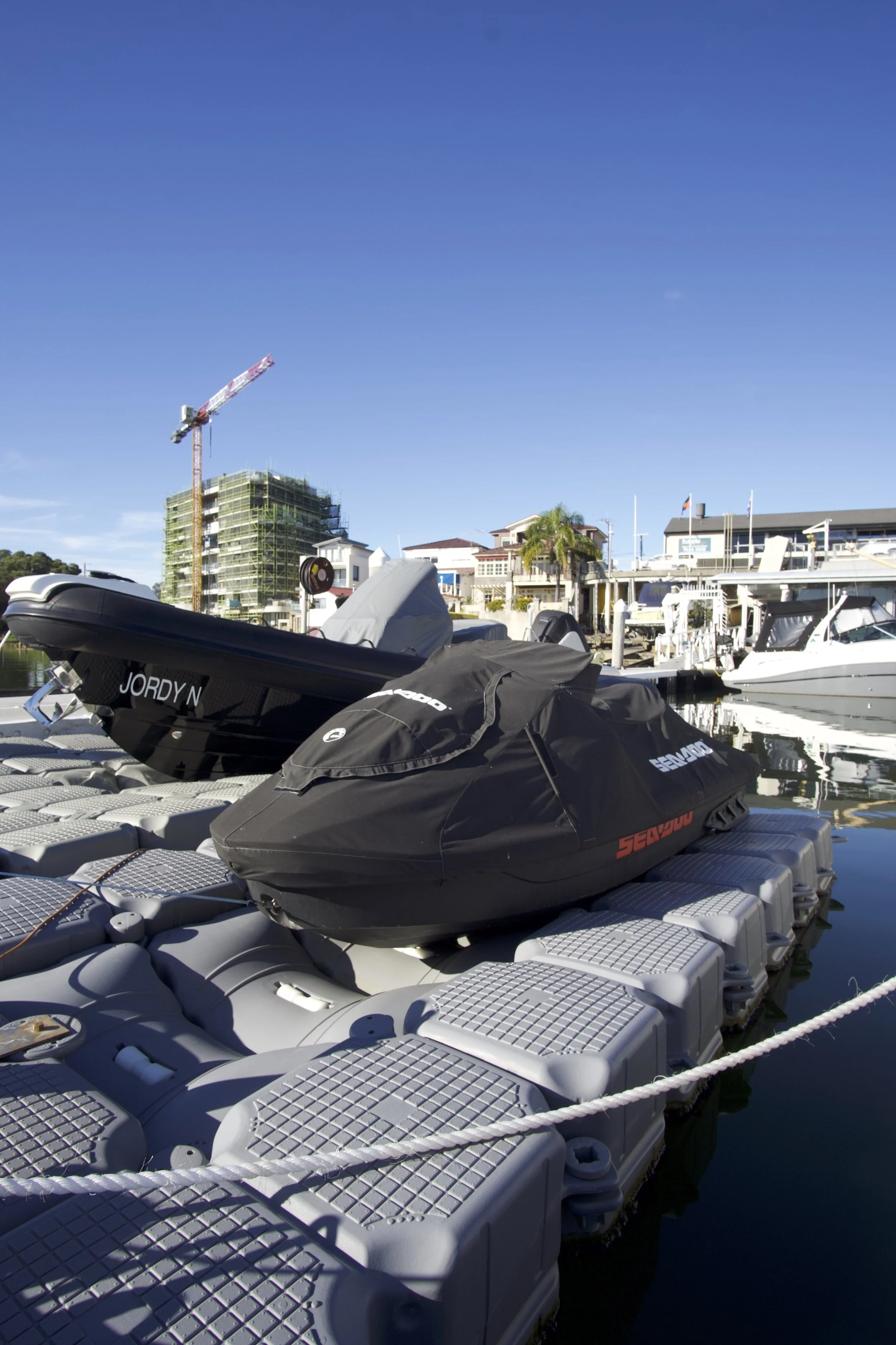 Boats docked at a marina on a clear, sunny day with blue sky, with residential buildings and construction crane in the background.