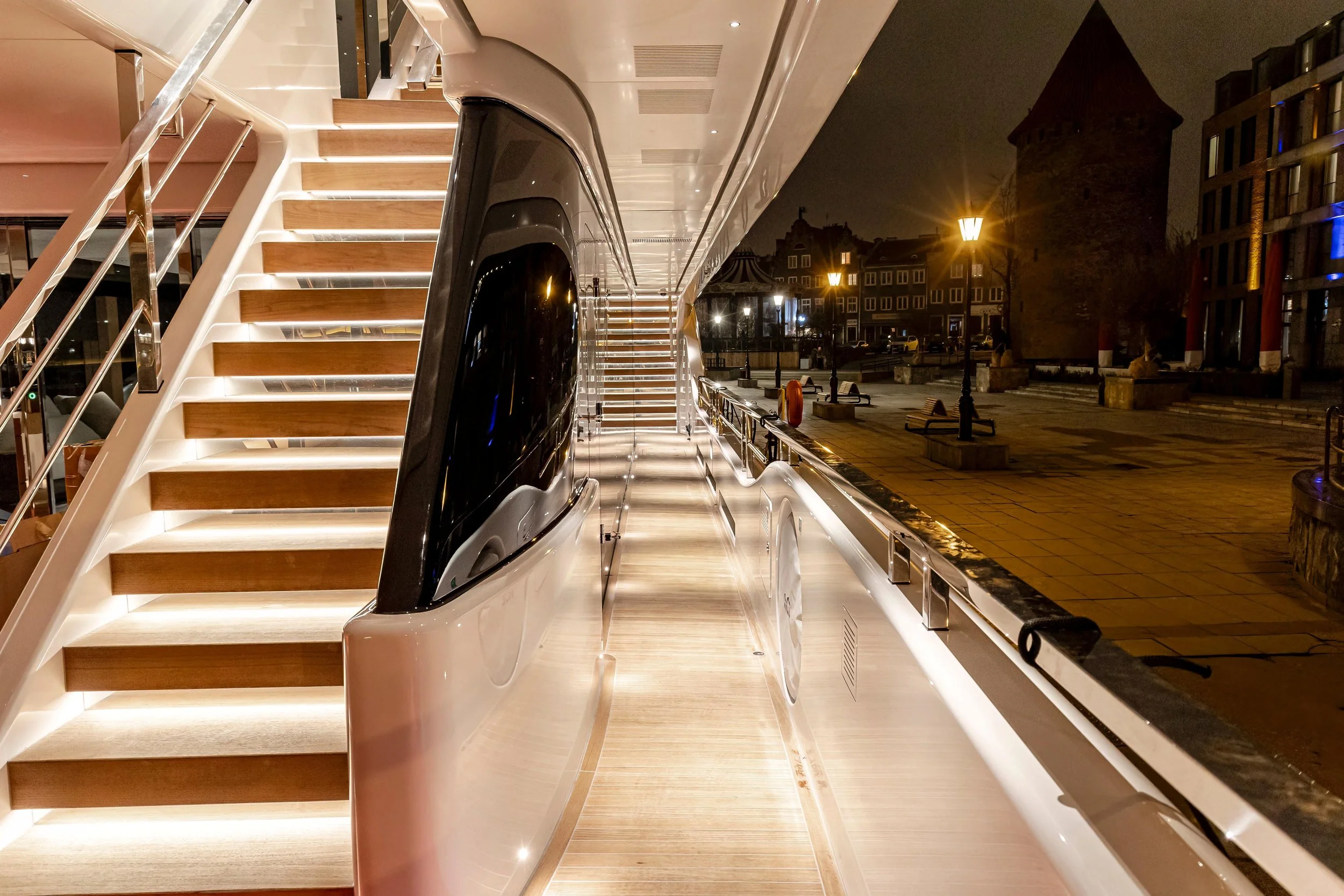 Nighttime view of the exterior side of a modern train or bus with illuminated stairs leading up to an upper deck, situated on a city street with lampposts, benches, and historic buildings visible in the background.