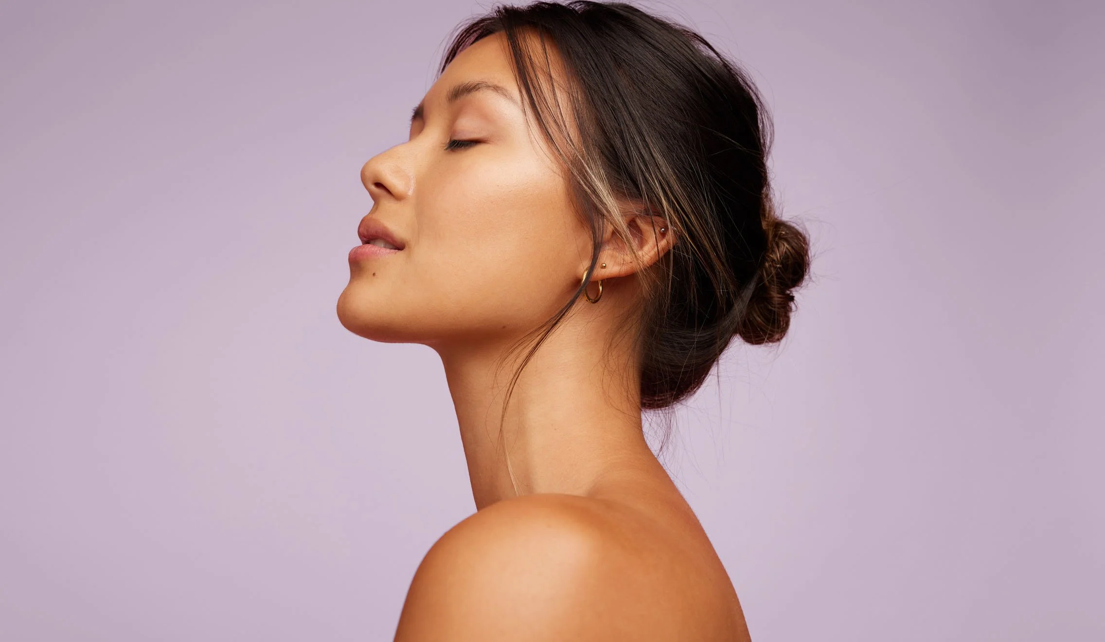 Side profile of a young woman with closed eyes, minimal makeup, and wearing small hoop earrings against a purple background.