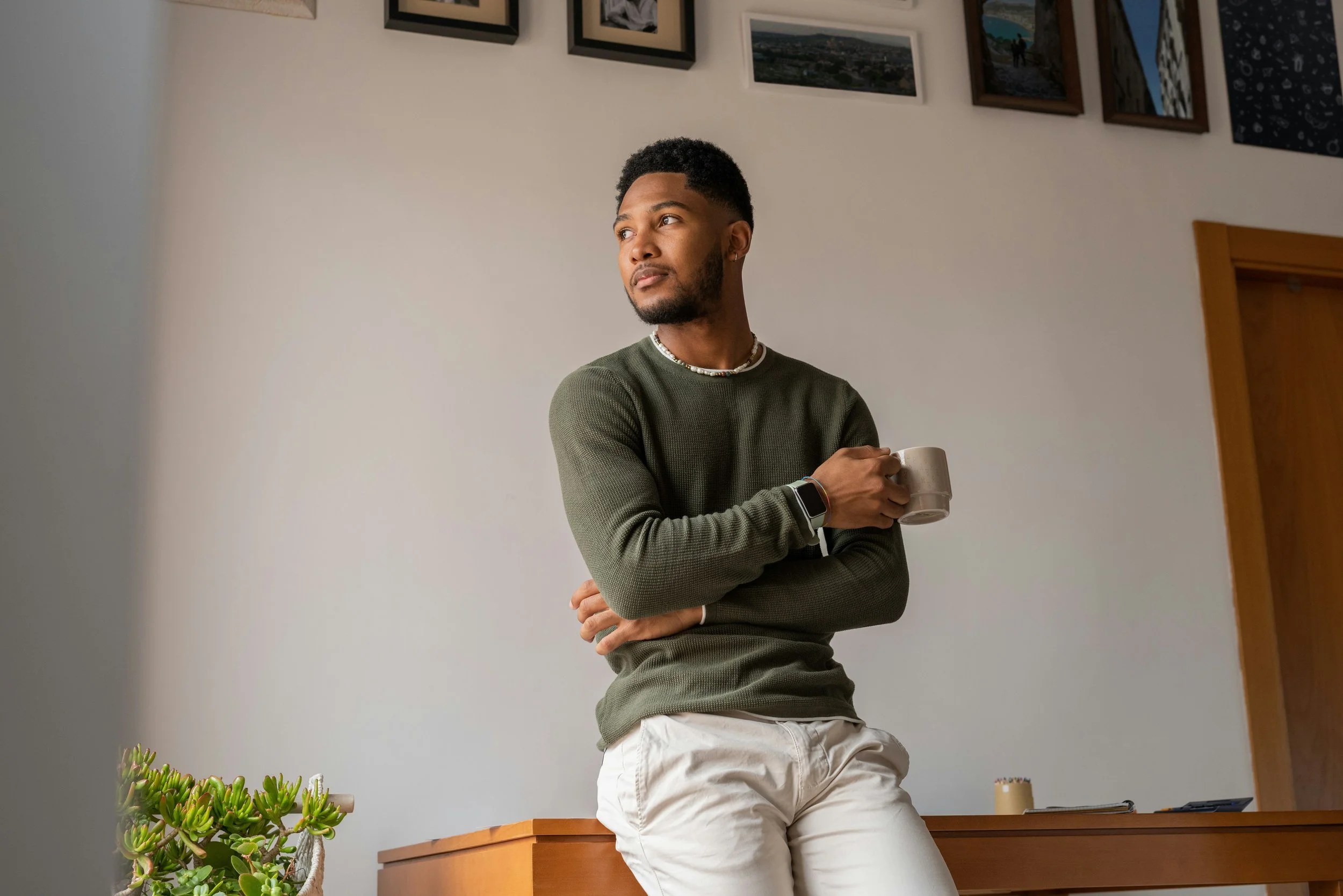 A man with a beard and short curly hair standing in front of a white wall with framed pictures, holding a mug, wearing a dark green sweater, white pants, and a smartwatch, with a plant and some desk items nearby.