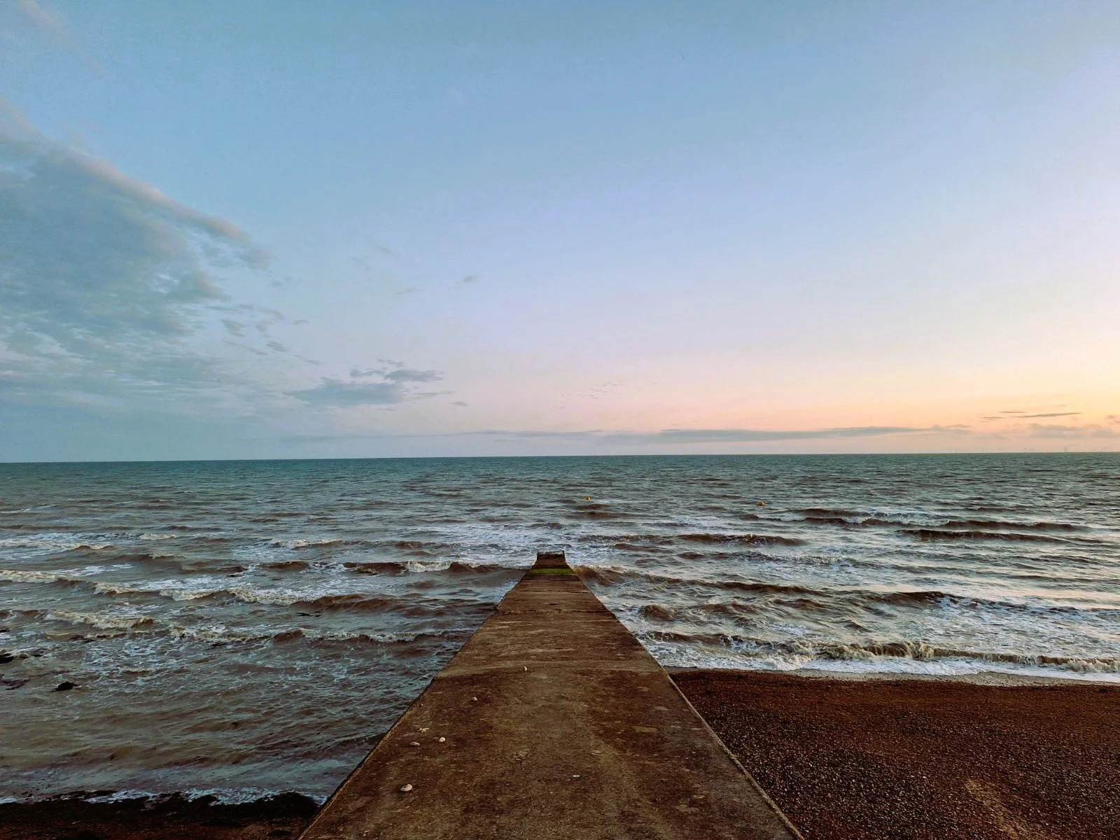 A concrete pier extending into the ocean with waves rolling onto the beach at sunset.