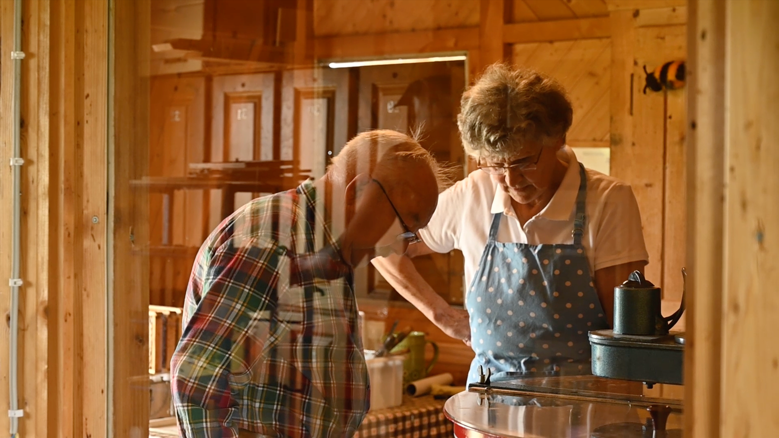 Ein älterer Mann und eine ältere Frau schauen gemeinsam durch eine Glasscheibe in eine warme Holz-Küche, in der sie zusammen arbeiten.