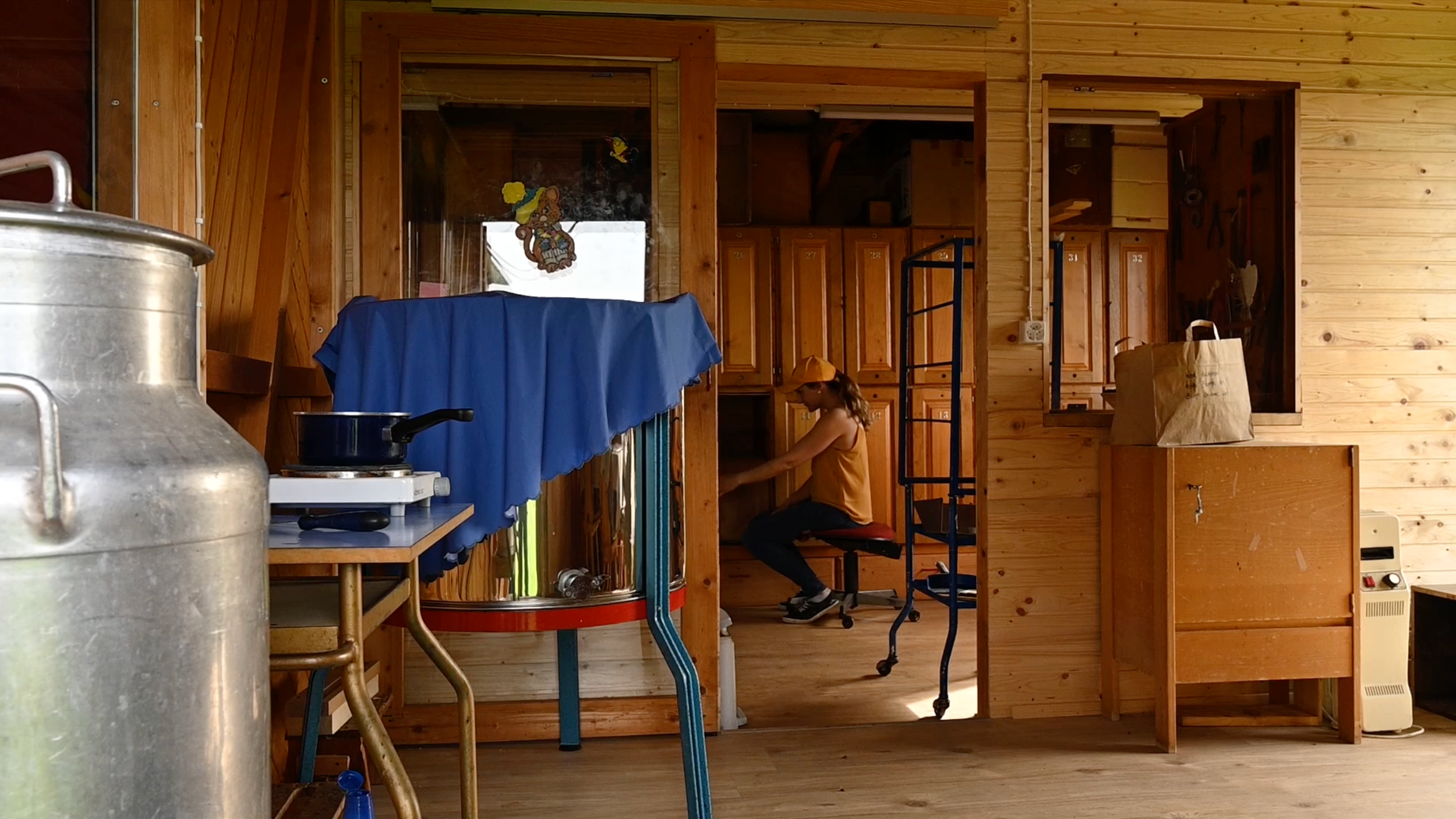 Junge Frau in einer Holzwerkstatt, die an einem Arbeitstisch arbeitet, mit einer Tacker, die im Hintergrund sichtbar ist, und weiteren Holzmöbeln und -kisten.