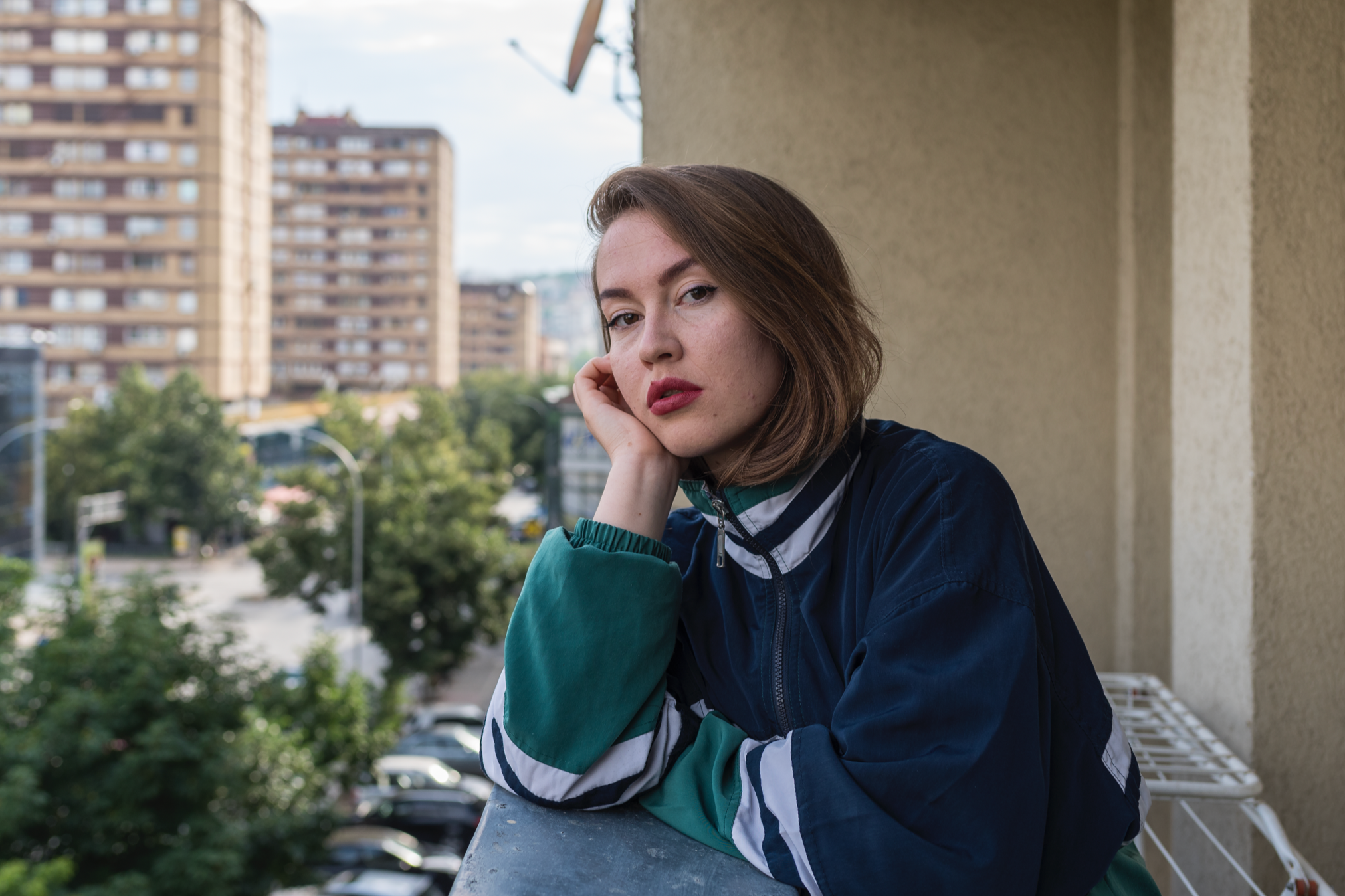 Junge Frau mit braunen Haaren auf einem Balkon, schauen nach vorne, trägt eine bunte Jacke, im Hintergrund ein Stadtbild mit Hochhäusern und Bäumen.