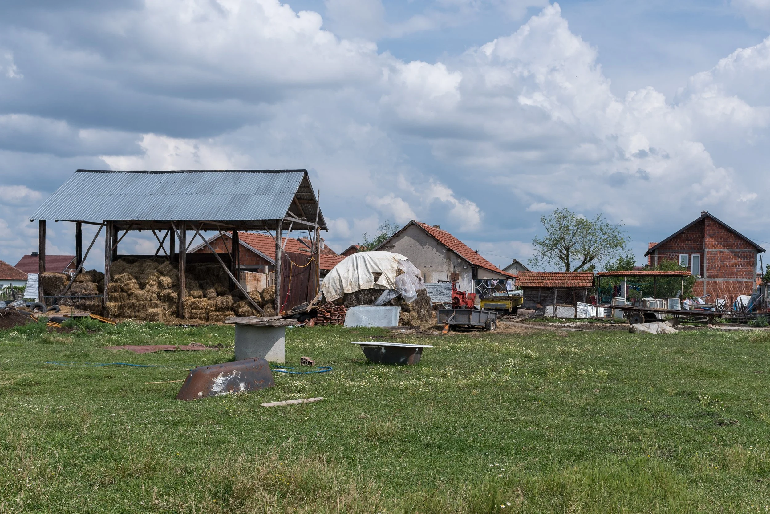 Bauernhof mit Lagerhäusern, Heuhaufen und landwirtschaftlichen Geräten unter einem bewölkten Himmel.