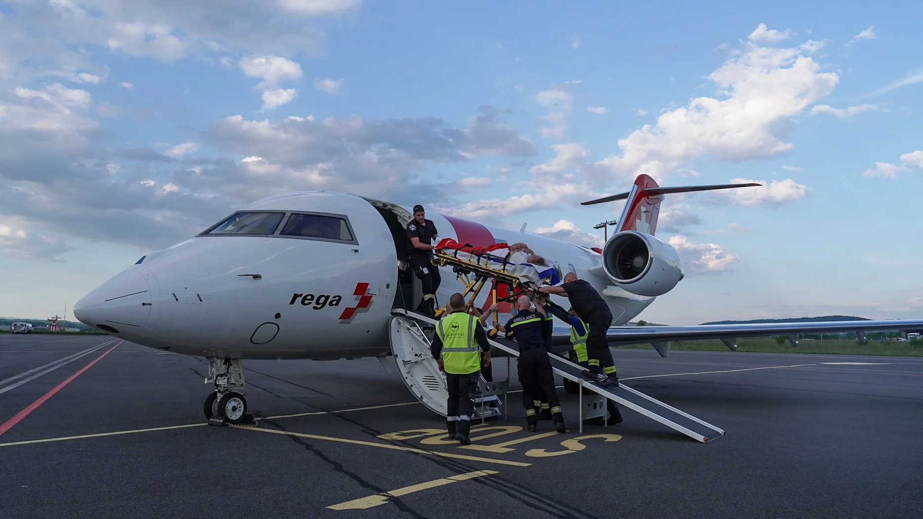 Ärzte und Sanitäter helfen einer Person auf einer Trage, die in einem Hubschrauber transportiert wird, auf einem Flugfeld bei Sonnenuntergang.